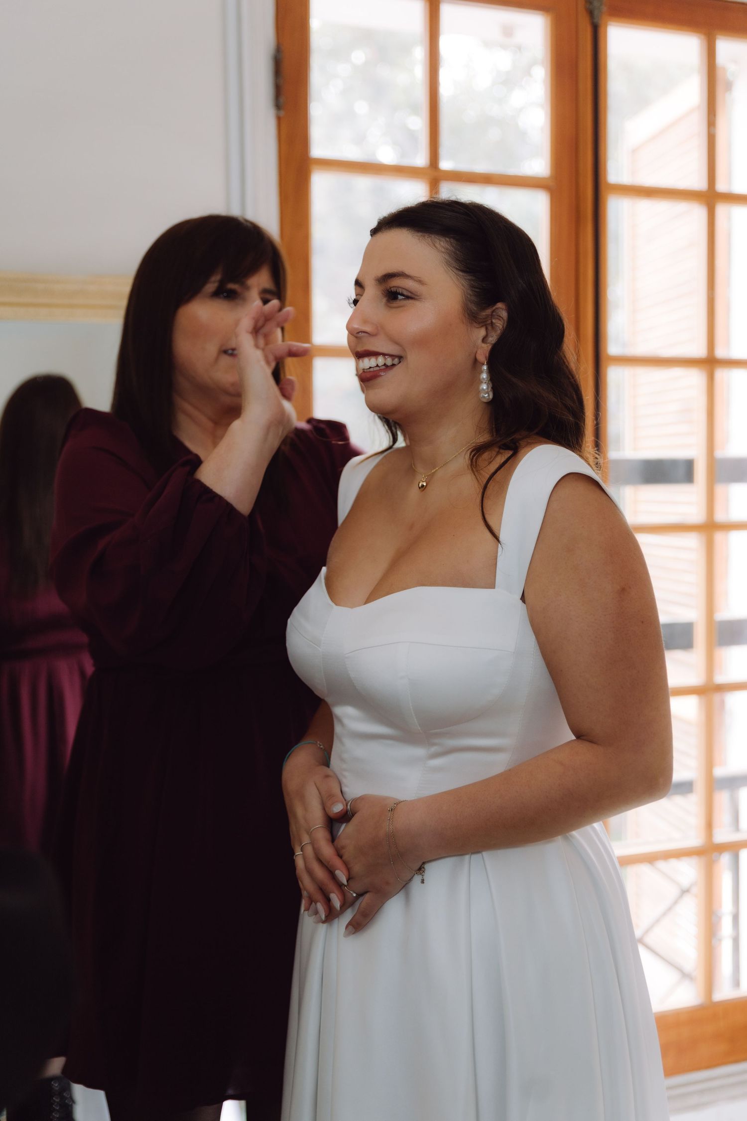 Makeup artist applies finishing touches to smiling bride in white dress near bright window.