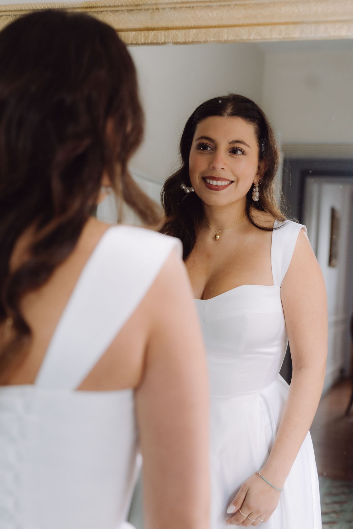 Radiant bride in white gown smiles at her reflection in elegant dressing room mirror.