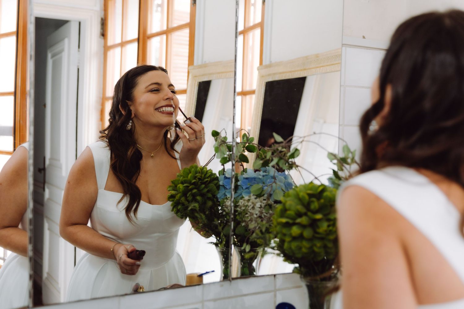 Bride applies makeup while smiling at mirror reflection surrounded by lush floral arrangements.