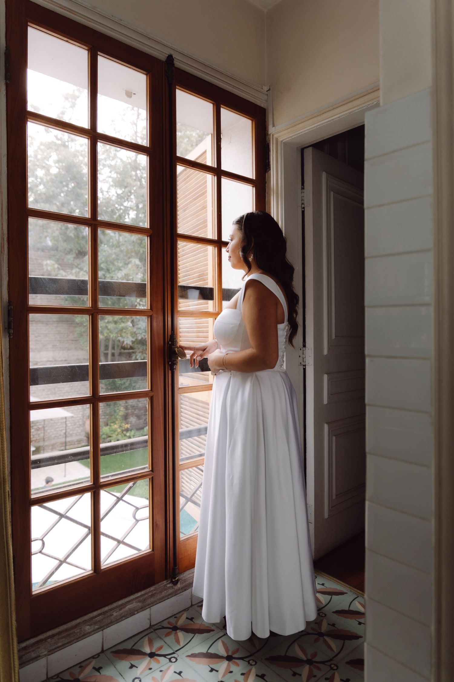 Bride in white gown gazing out ornate wooden French doors in elegant room with decorative tile floor.