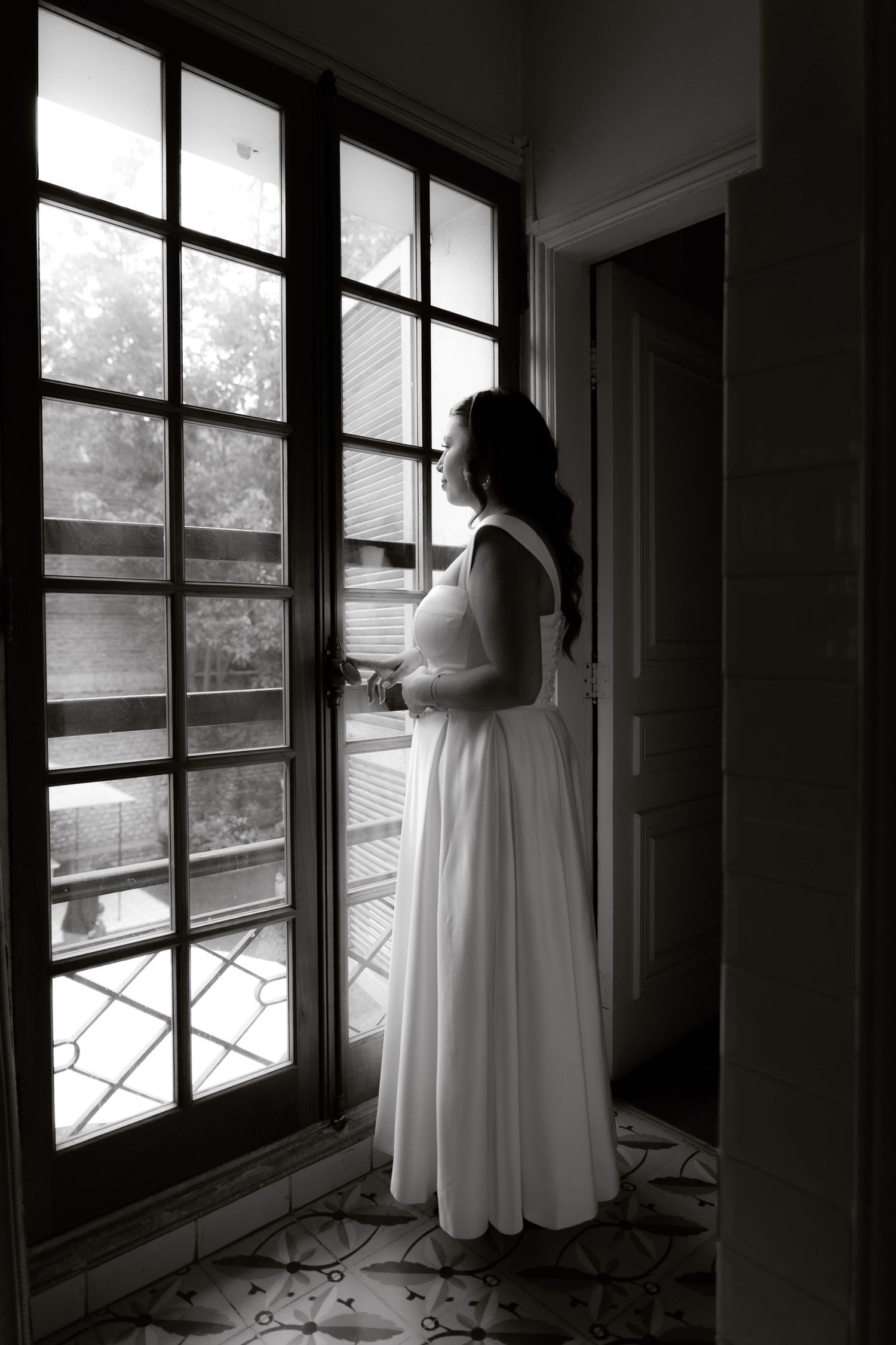 Black and white photo of bride in white dress standing by French windows in contemplative pose.