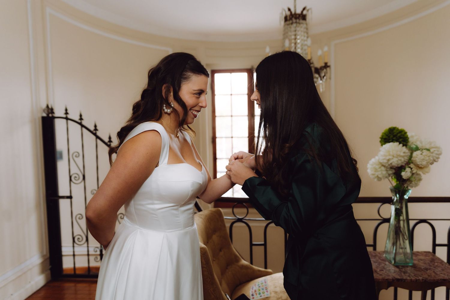 Bride in white wedding gown laughing joyfully while bridesmaid in green dress fastens bracelet.