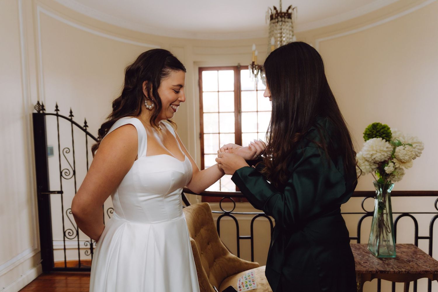 Bride smiling warmly as bridesmaid in dark green dress helps her put on jewelry before wedding.