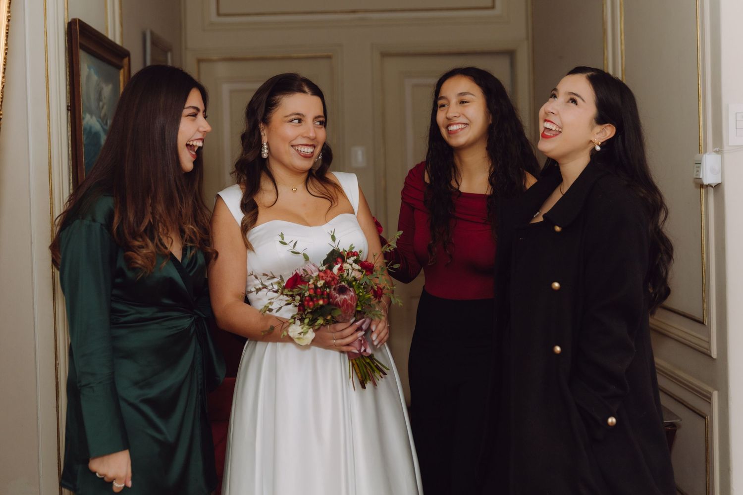 Bride holding red bouquet laughing with three bridesmaids in elegant hallway before wedding ceremony.