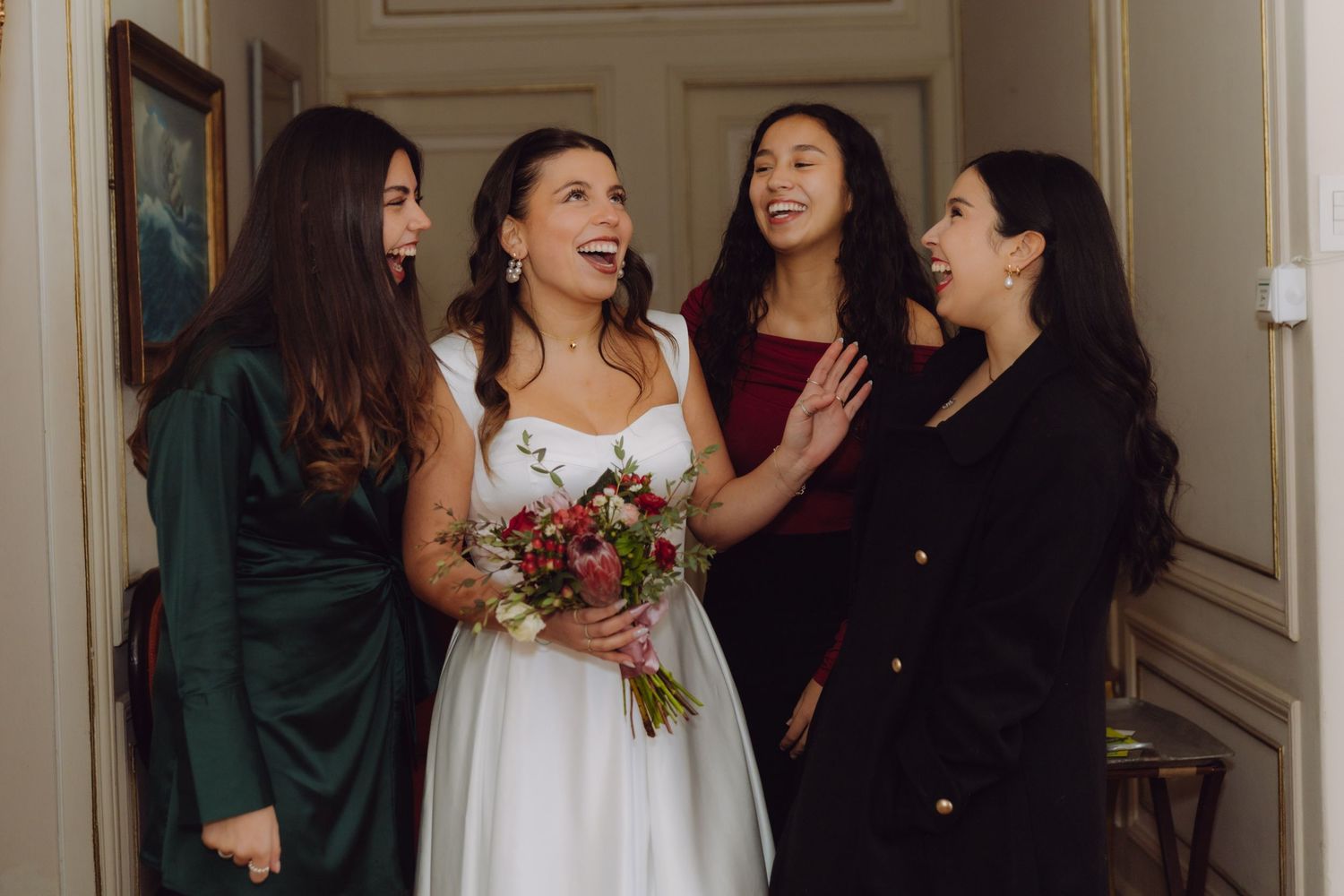 Radiant bride with red bouquet sharing joyful moment with three bridesmaids in classic interior hallway.