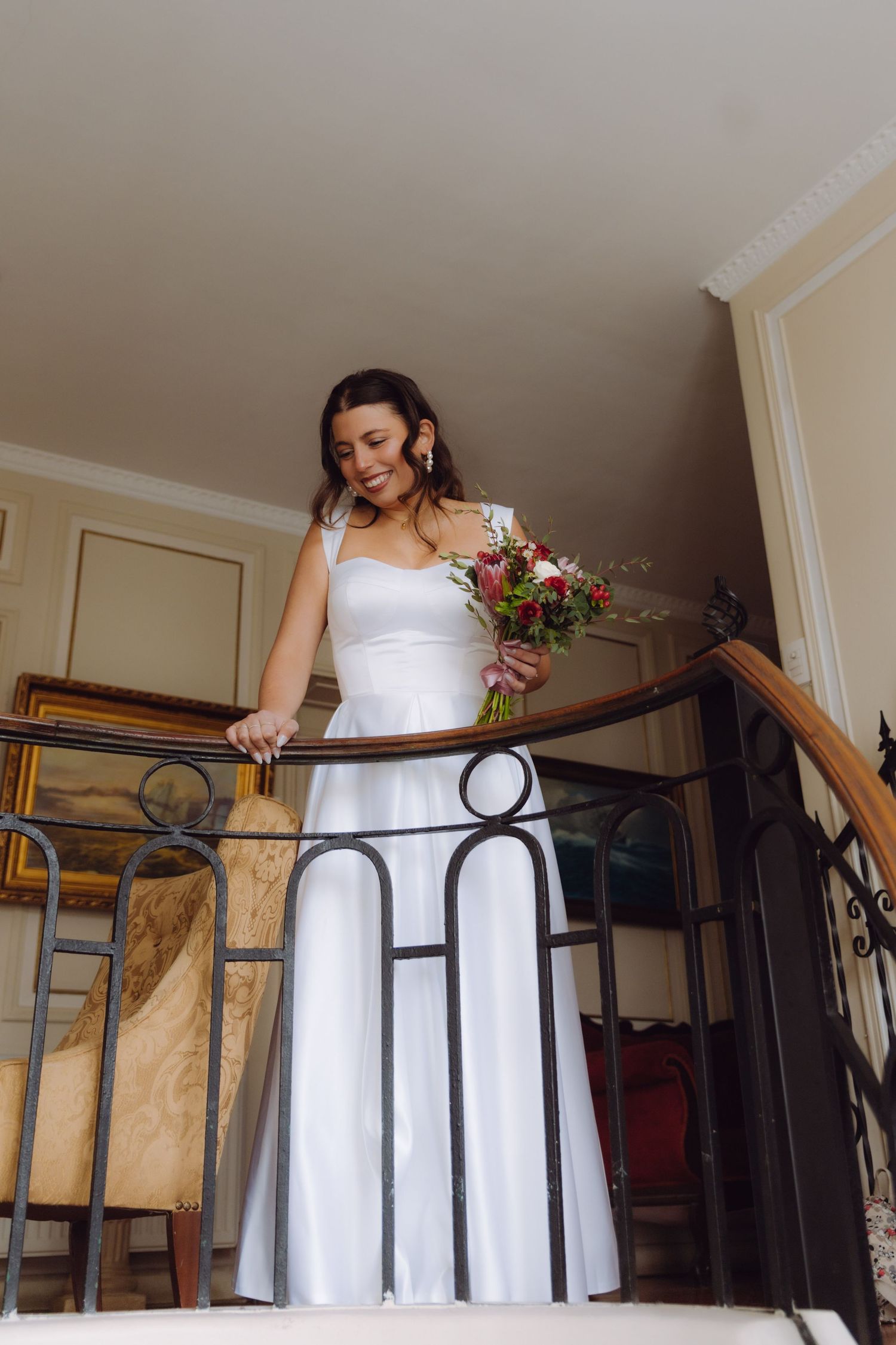 Elegant bride in white gown holding red bouquet descending ornate wrought iron staircase indoors.