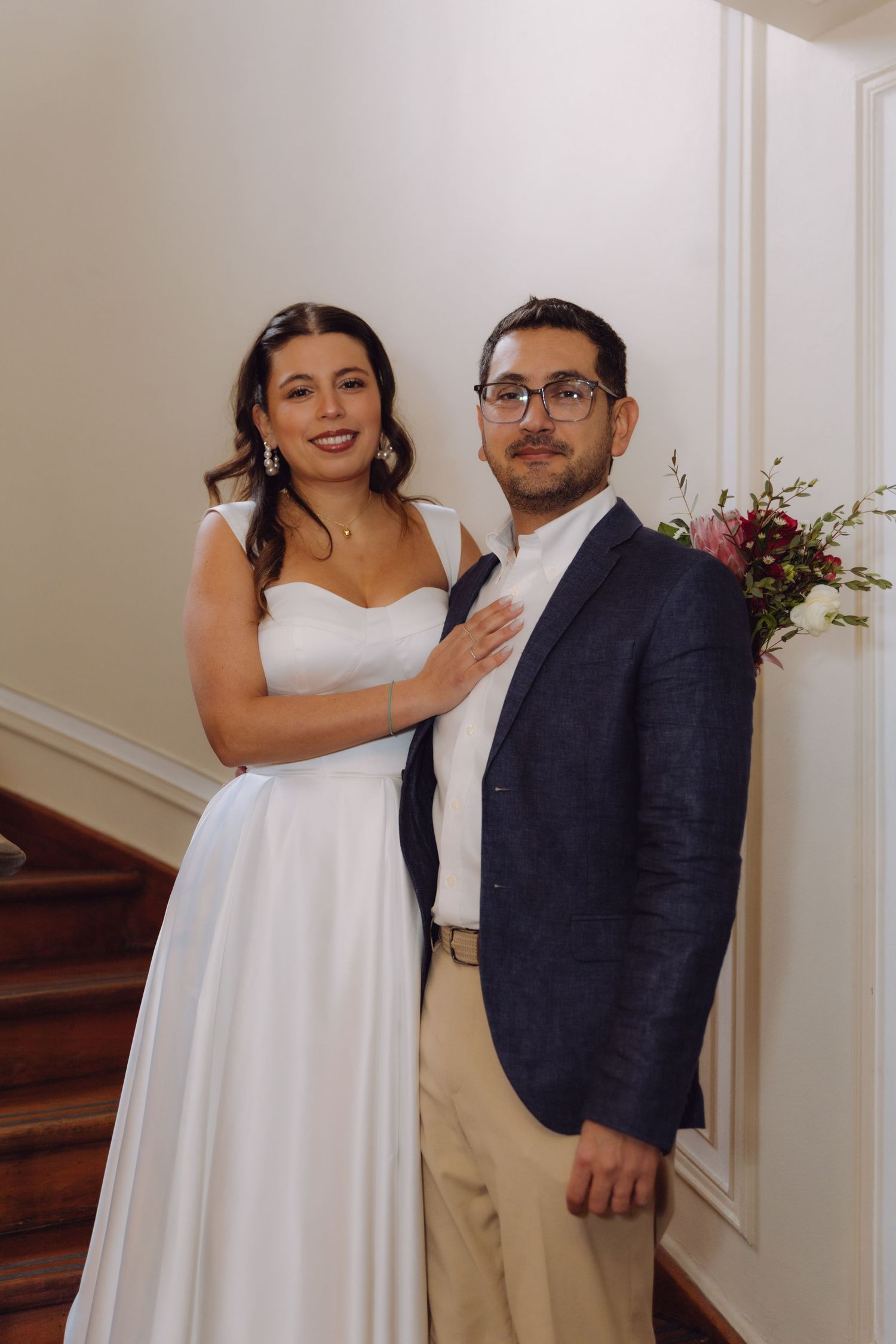 Bride in white gown and groom in navy blazer posing together near staircase with floral arrangement.