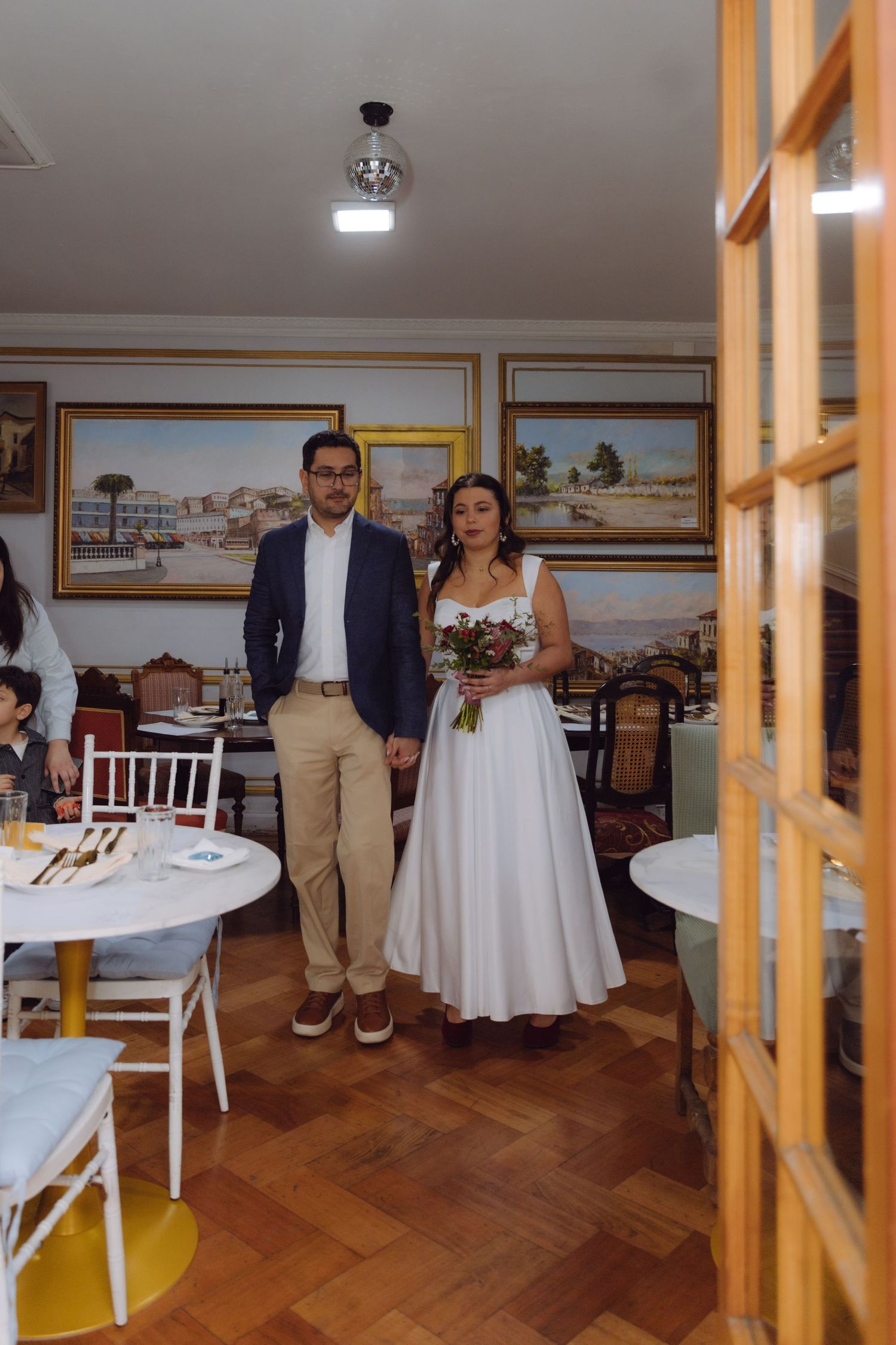 Bride and groom holding hands walking through dining room with landscape paintings on walls.