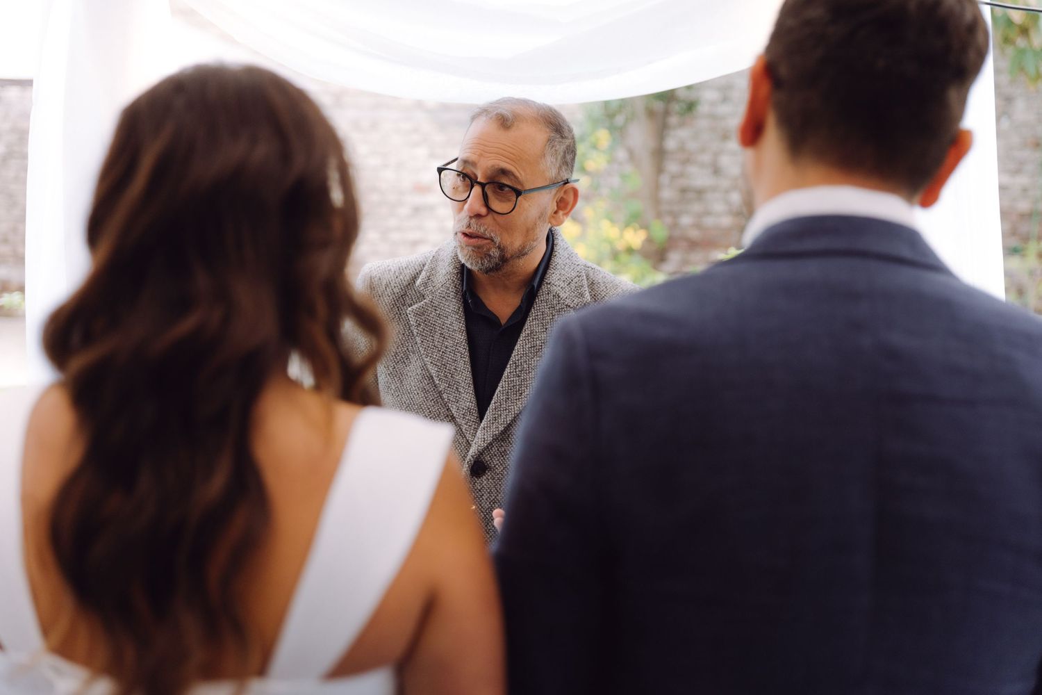 Wedding officiant in gray jacket and glasses faces couple during intimate outdoor ceremony.