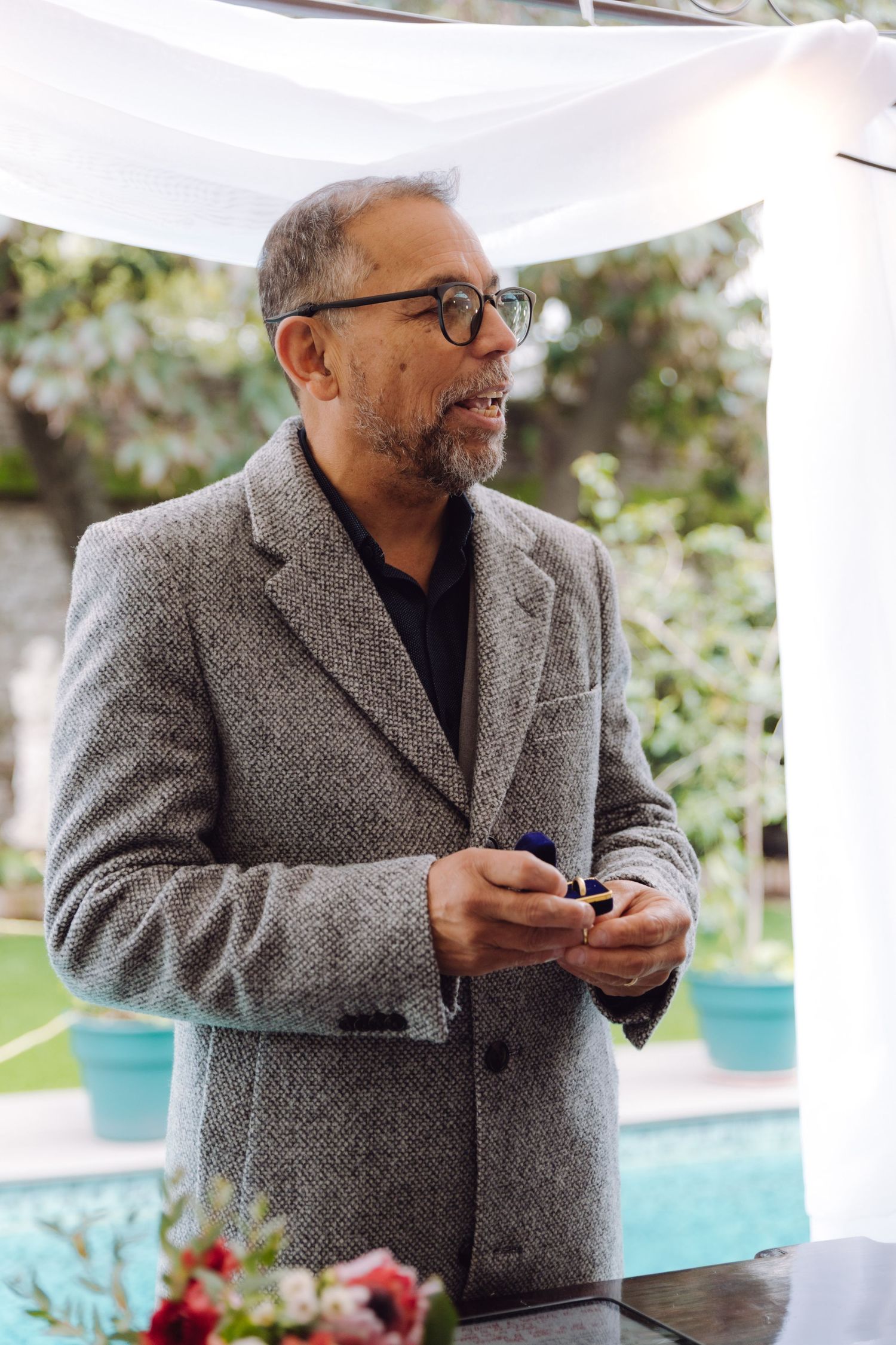 Mature man in gray tweed jacket holding wedding rings at outdoor ceremony altar.