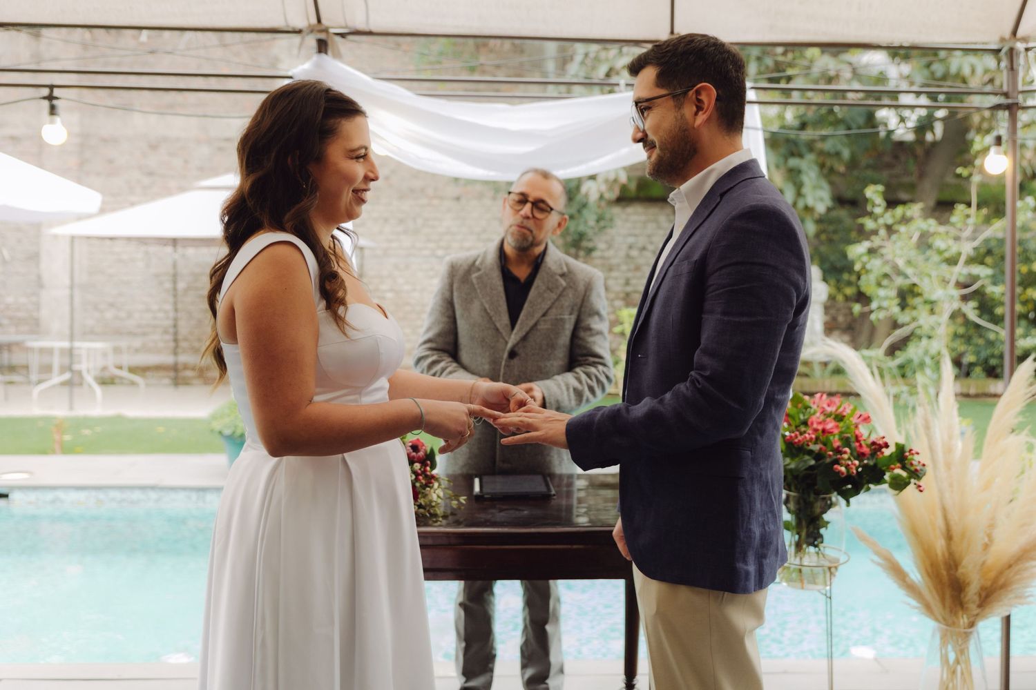 Smiling bride and groom exchange wedding rings with officiant behind them at outdoor ceremony.