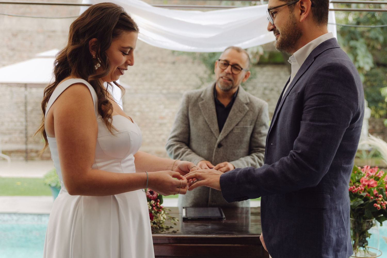 Bride smiles as groom places wedding ring on her finger during outdoor pool ceremony.