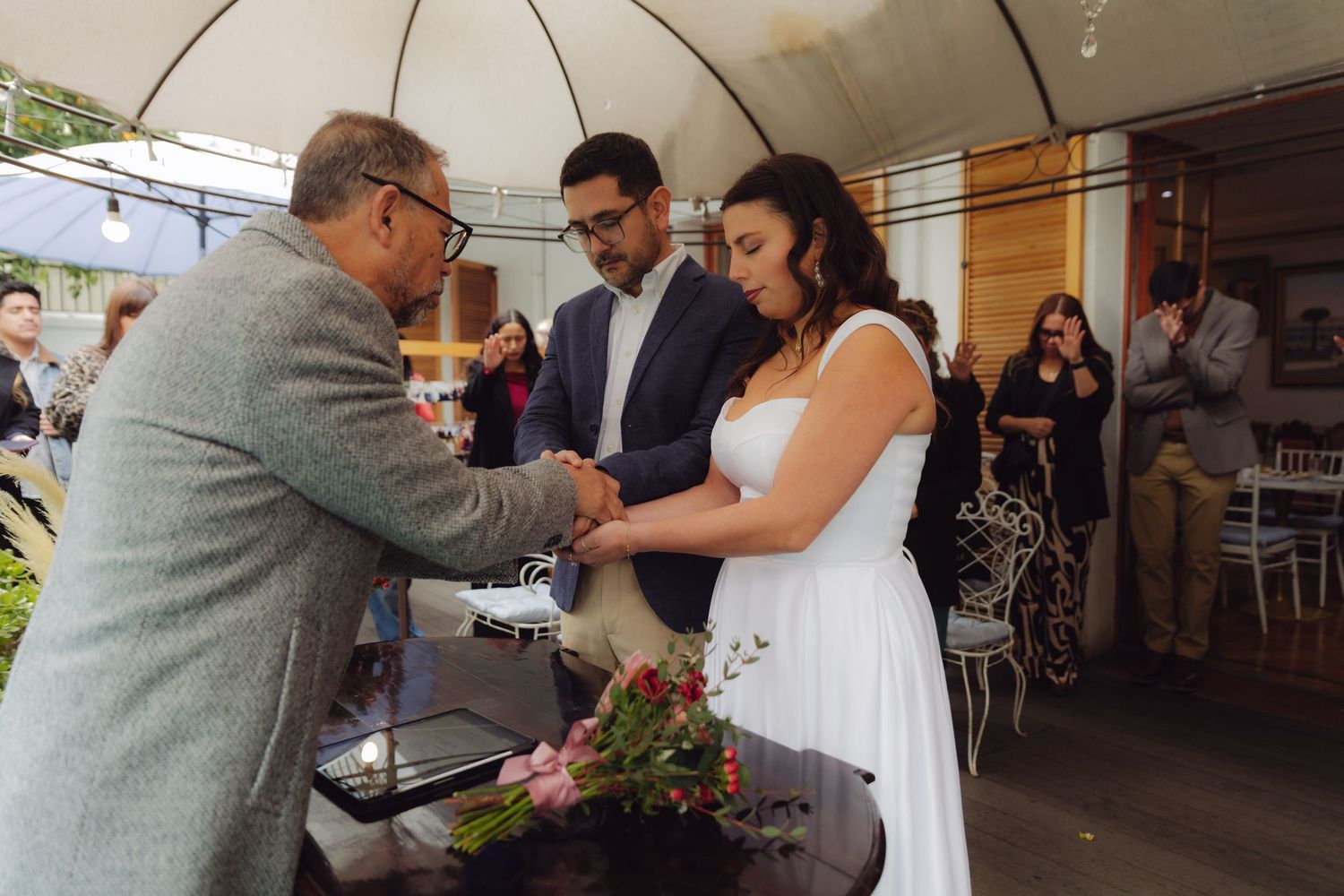 Wedding officiant shakes hands with couple at flower-adorned ceremony table with guests watching.