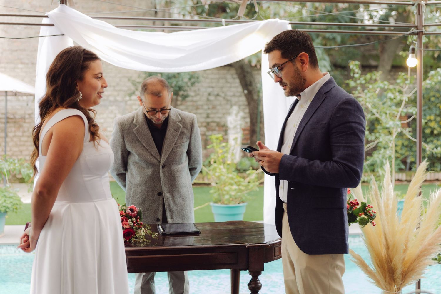 Groom reading vows from phone at elegant outdoor poolside wedding ceremony.