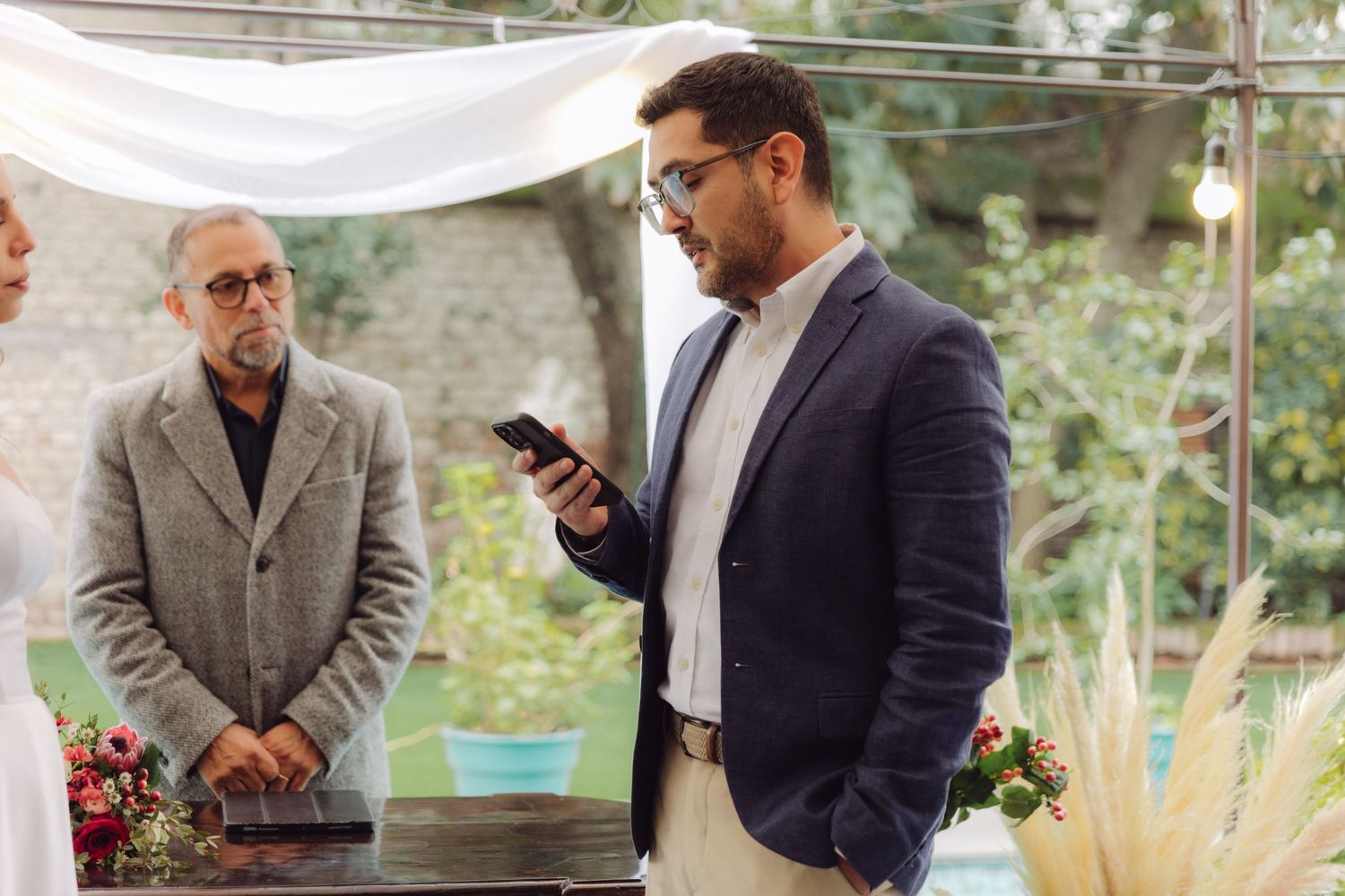 Groom reading wedding vows from smartphone at elegant outdoor ceremony.