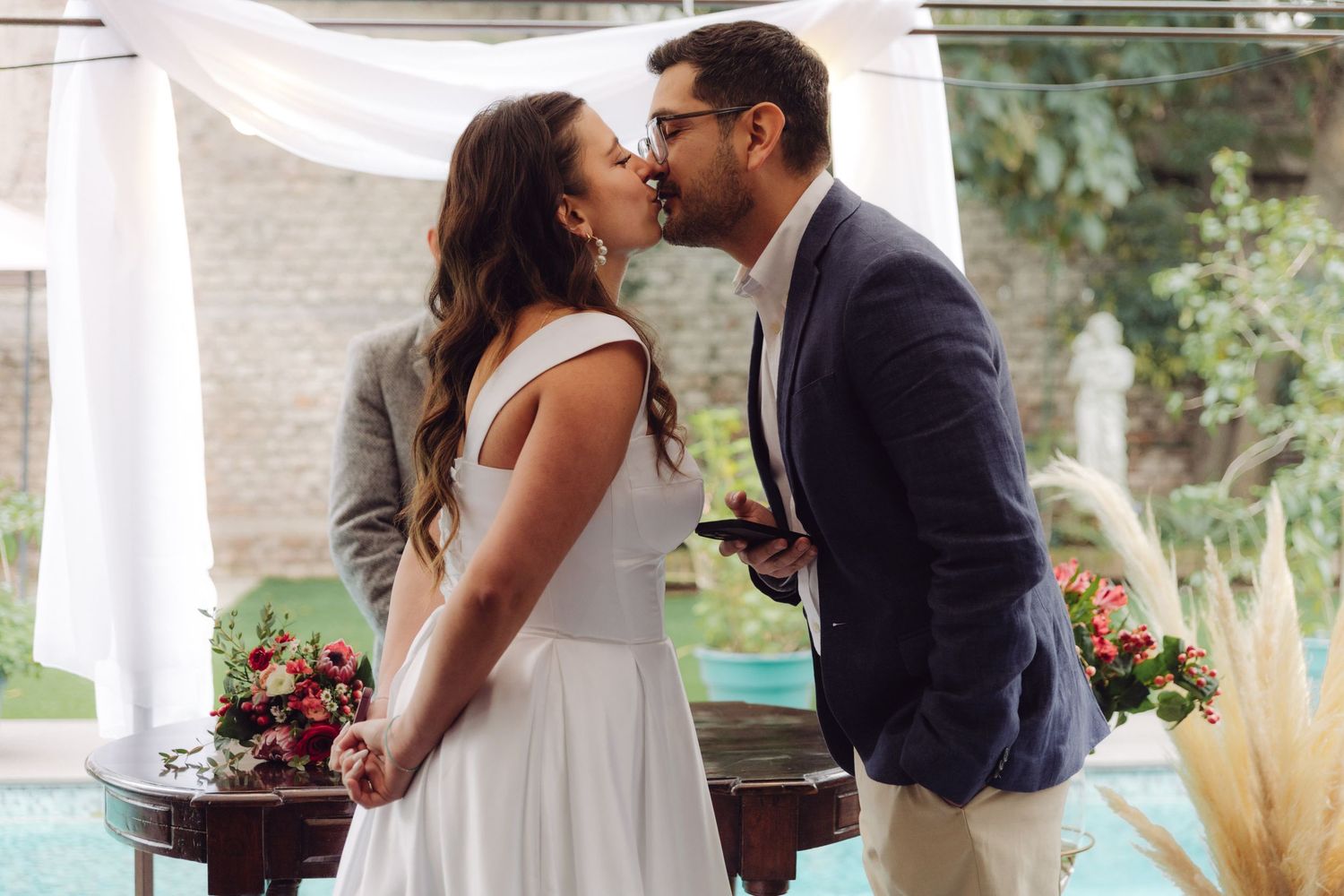 Bride and groom share a kiss at outdoor wedding ceremony near pool with floral decorations.