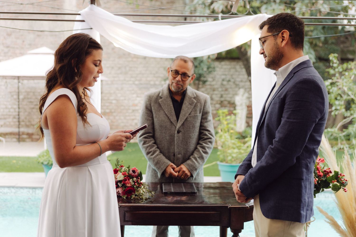 Bride reads vows while officiant and groom stand at outdoor wedding altar near pool.