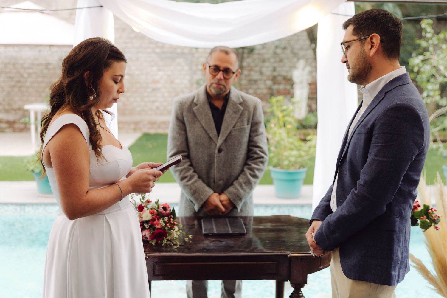 Bride holds bouquet reading vows as officiant and groom watch at outdoor wedding ceremony.