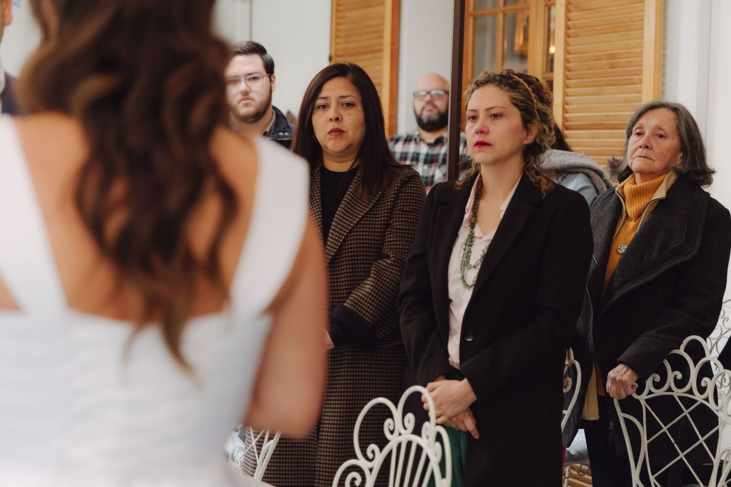 Wedding guests in formal attire watch bride during intimate indoor wedding ceremony.
