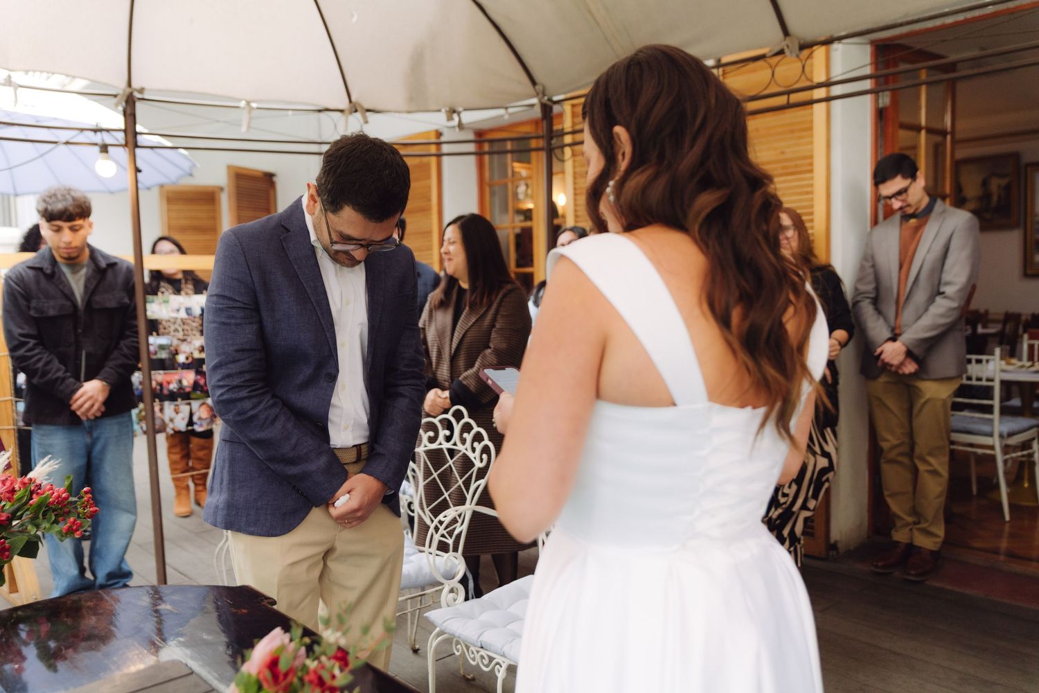 Groom bows head during wedding ceremony prayer while guests and bride stand nearby.