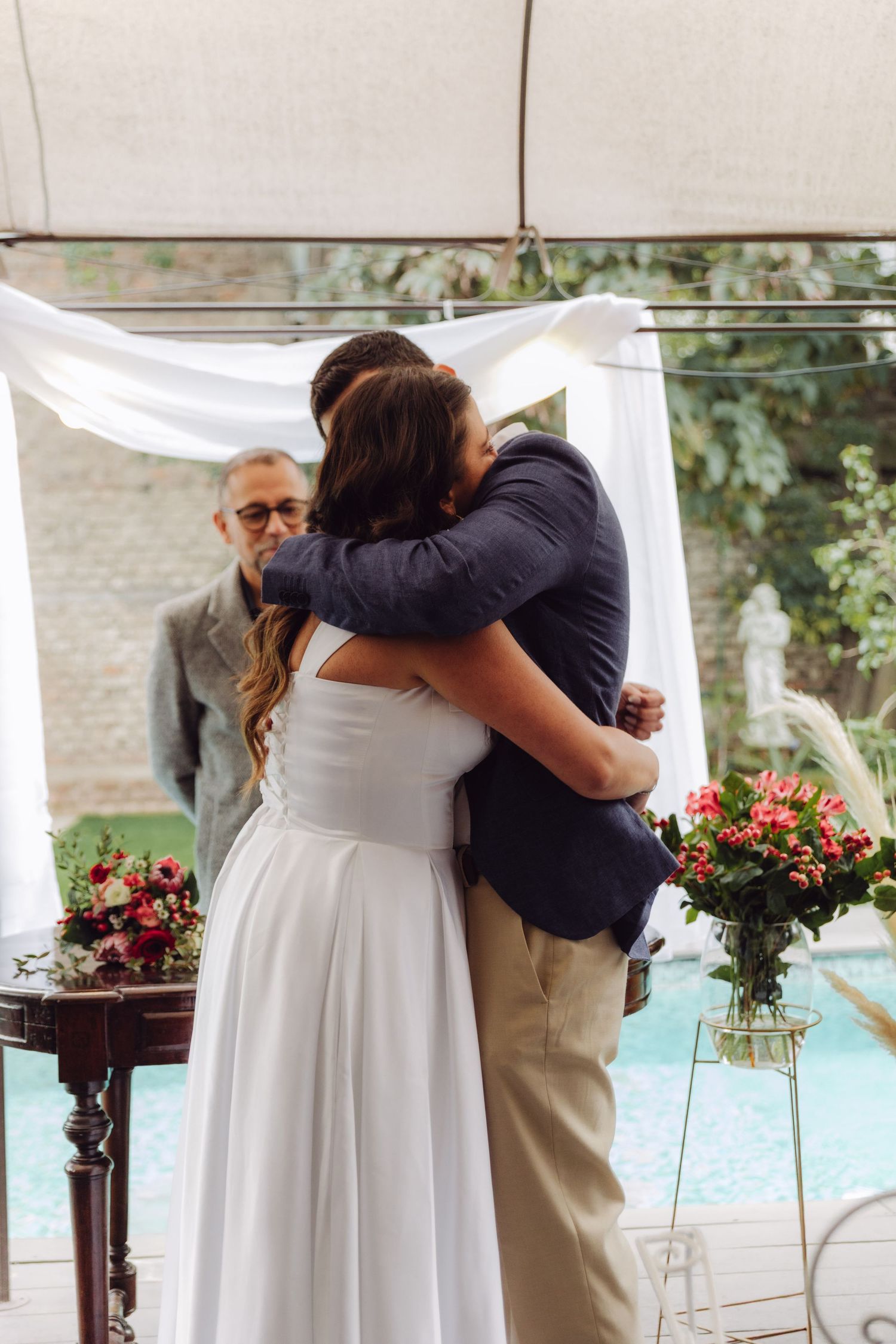 Newlyweds share heartfelt embrace at outdoor wedding altar as officiant watches behind them.