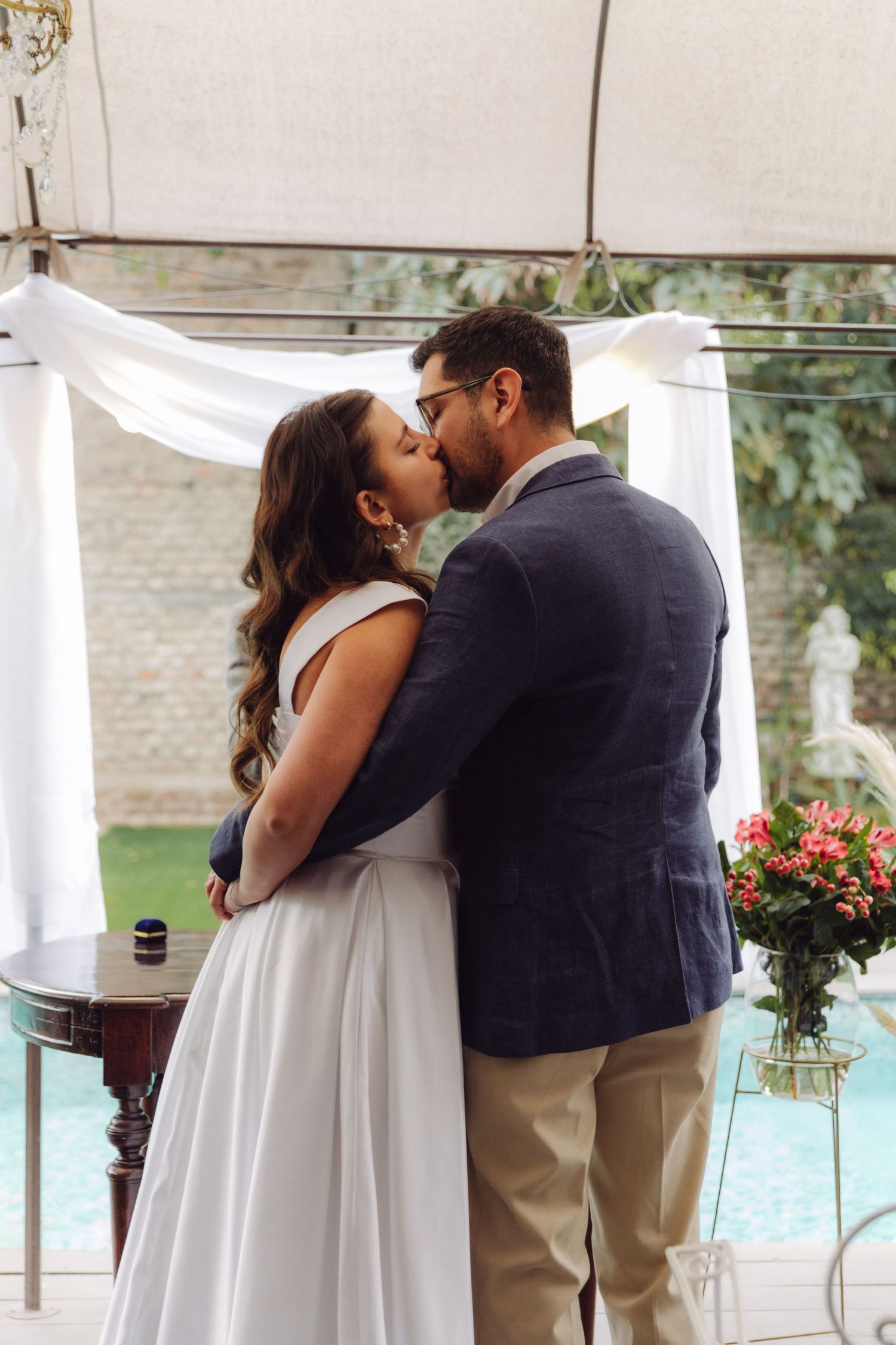 Bride and groom share romantic kiss at outdoor wedding ceremony beneath white draped arch.