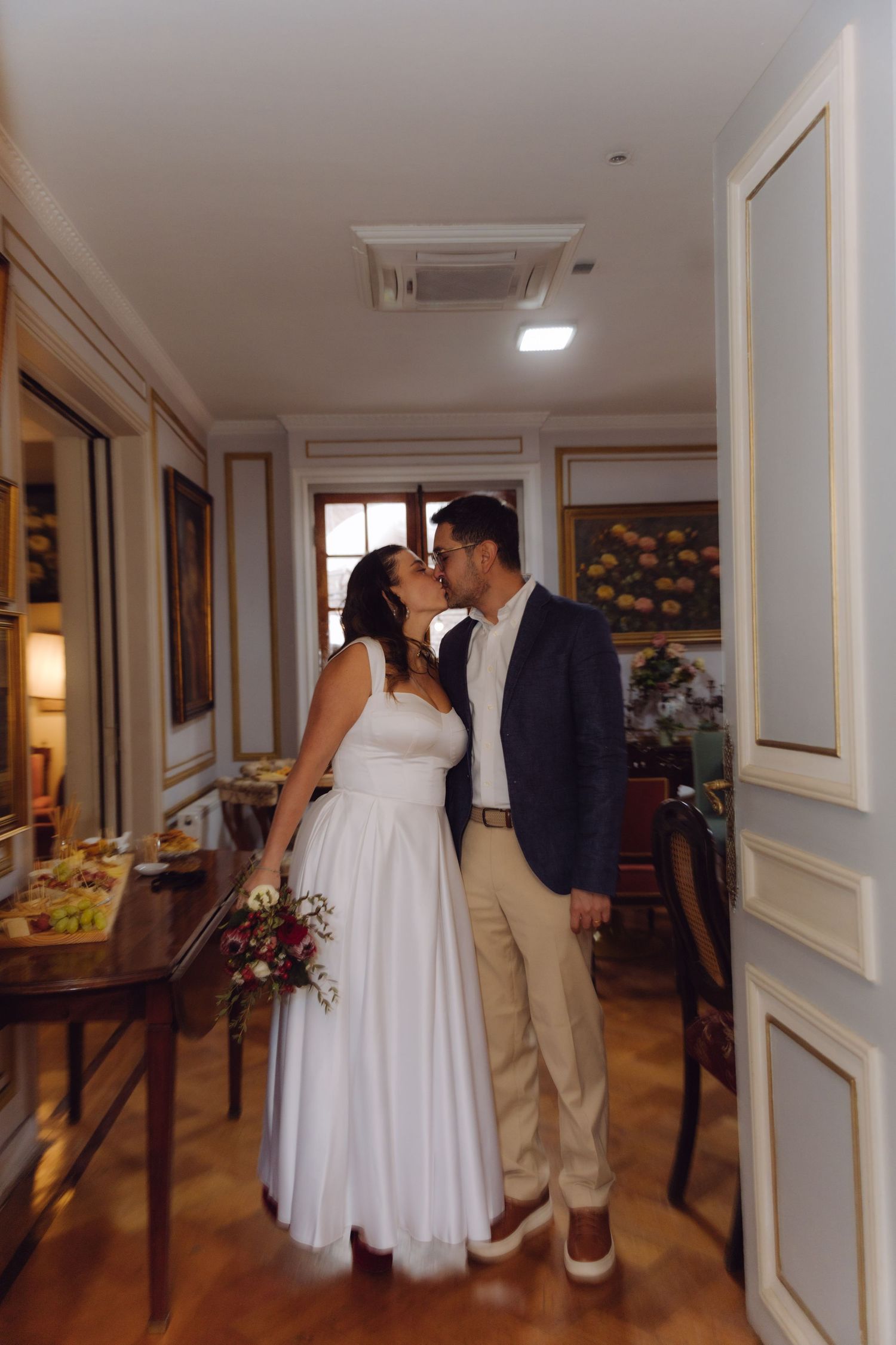 Newlyweds share a tender kiss inside elegant room while bride holds floral bouquet.
