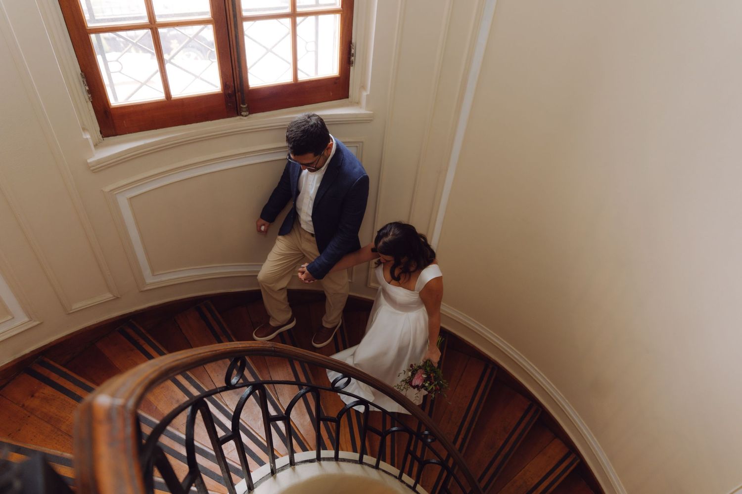 Newlyweds descend elegant spiral staircase together after indoor wedding ceremony.