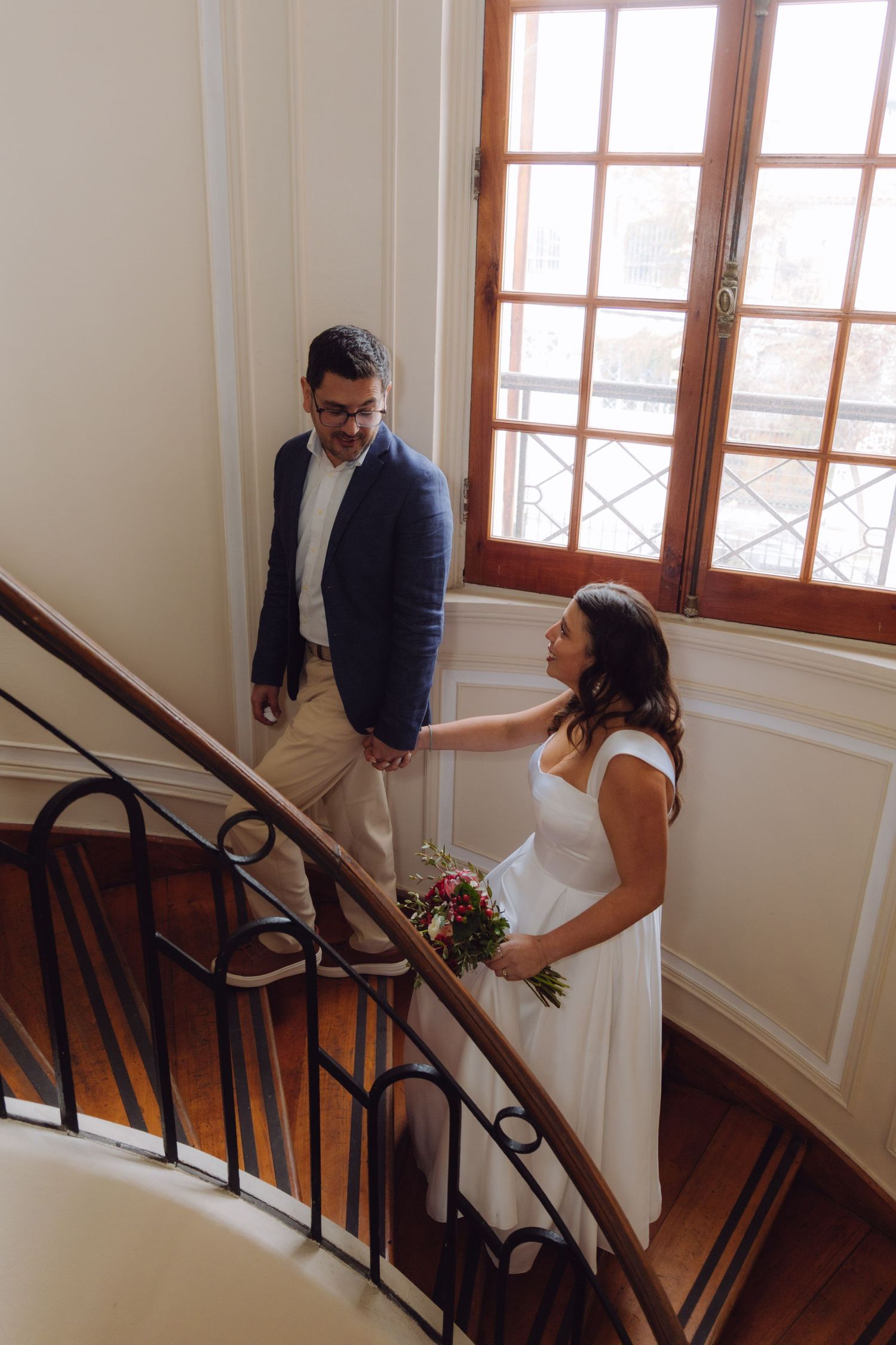 Bride in white dress holds bouquet on elegant staircase as groom in navy blazer smiles at her.