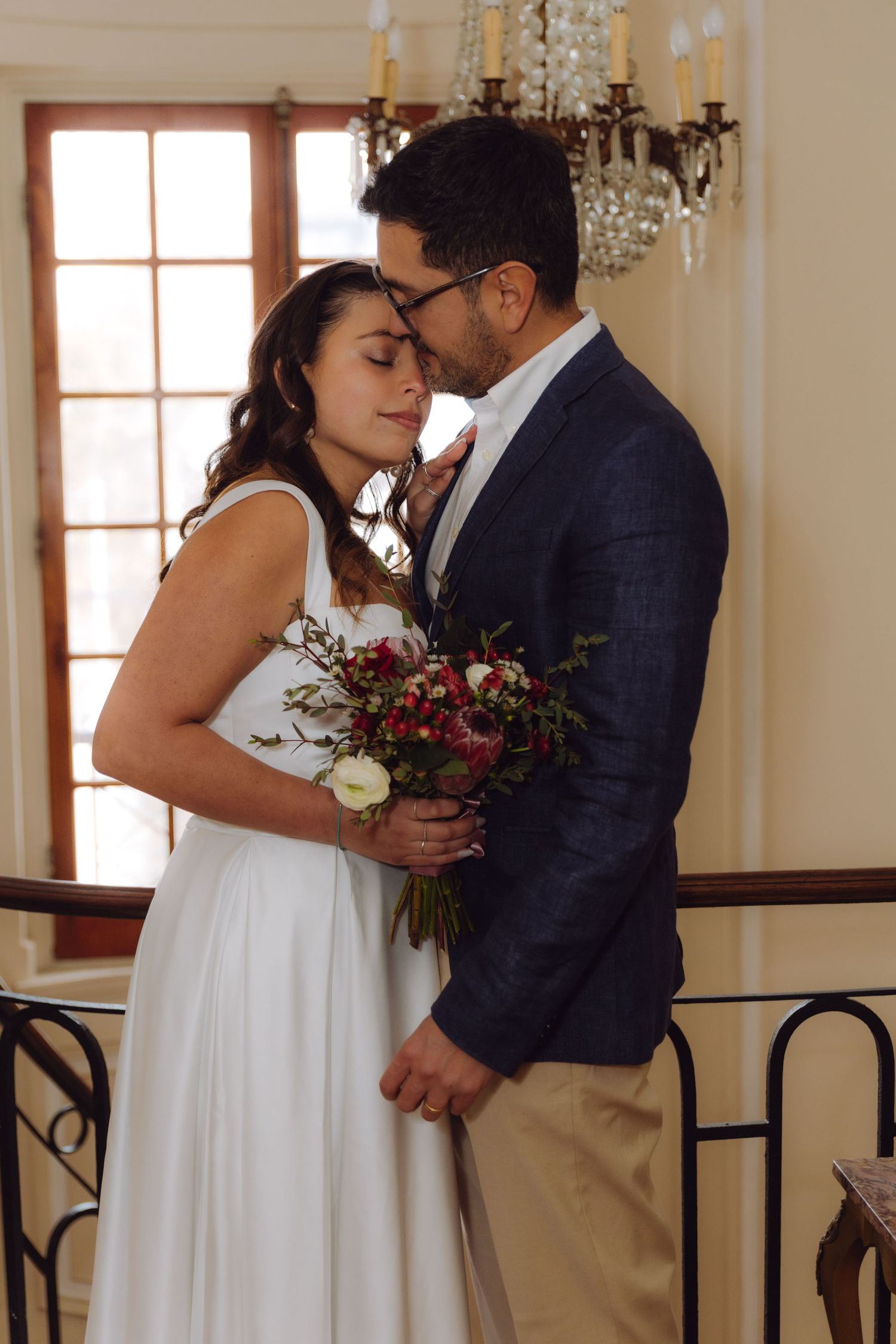 Groom tenderly kisses bride on forehead as she holds a lush red and white wedding bouquet.