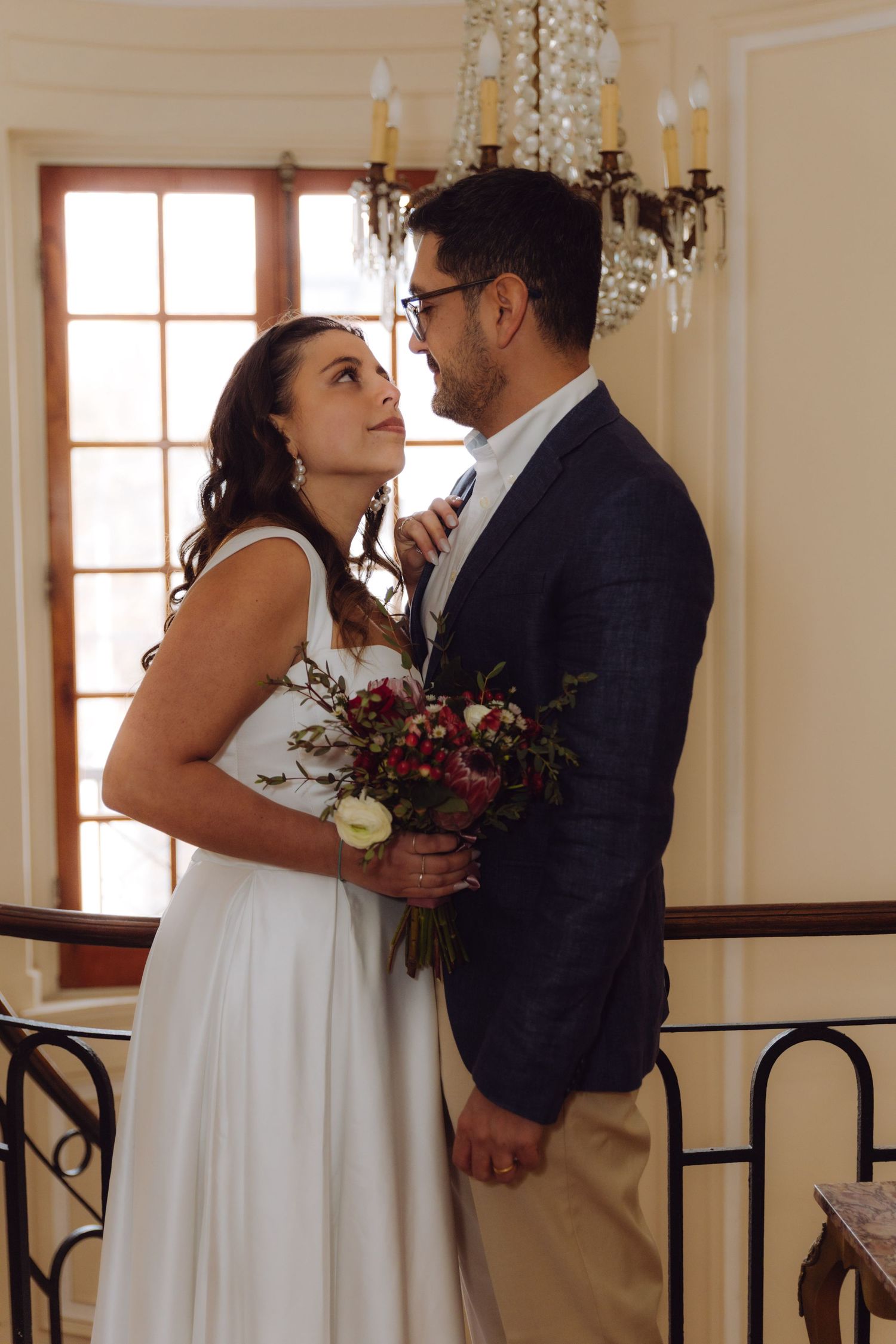 Wedding couple embraces near ornate chandelier, bride holding floral bouquet gazes up at groom.