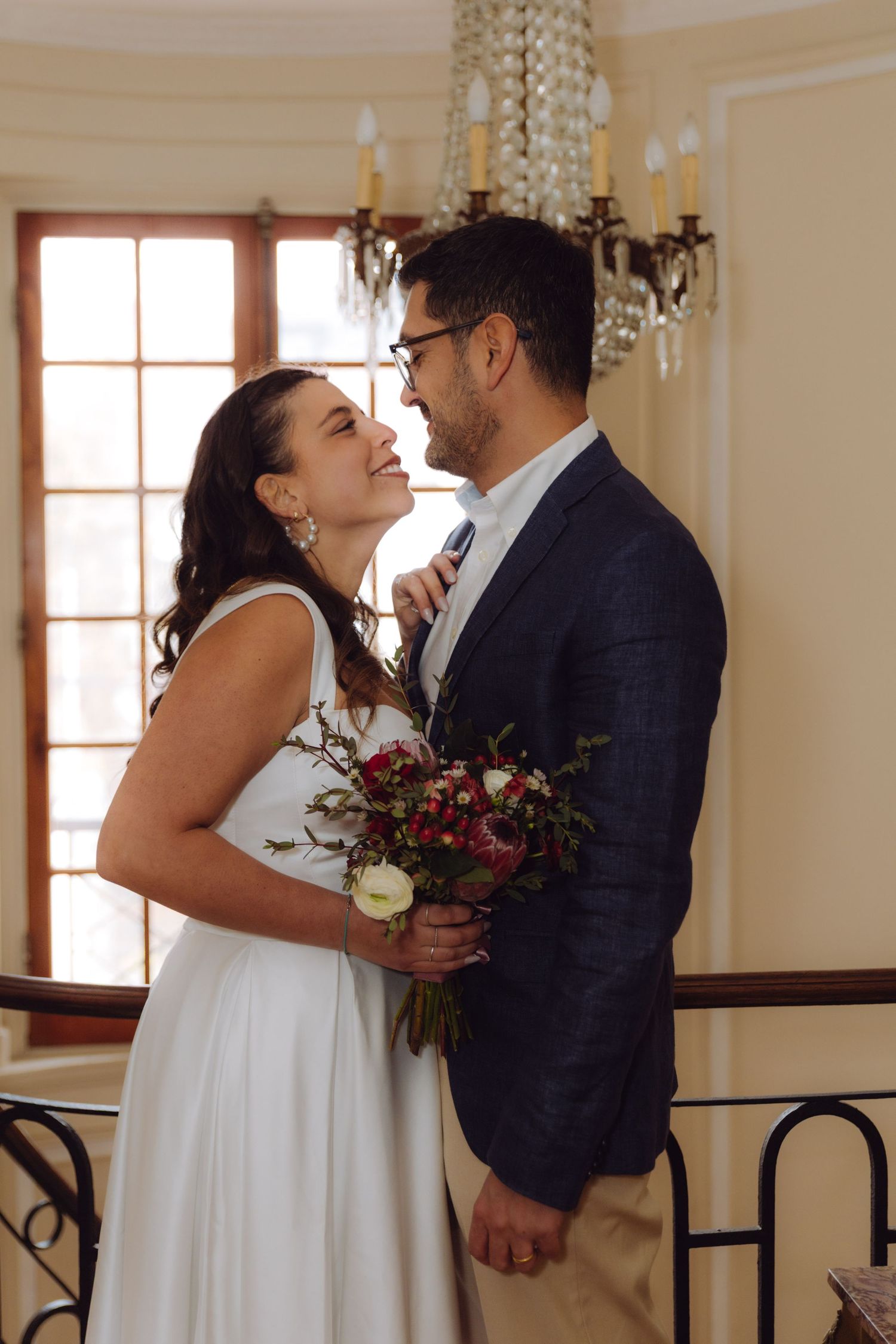 Smiling bride in off-shoulder white gown and groom in navy blazer share a romantic moment indoors.