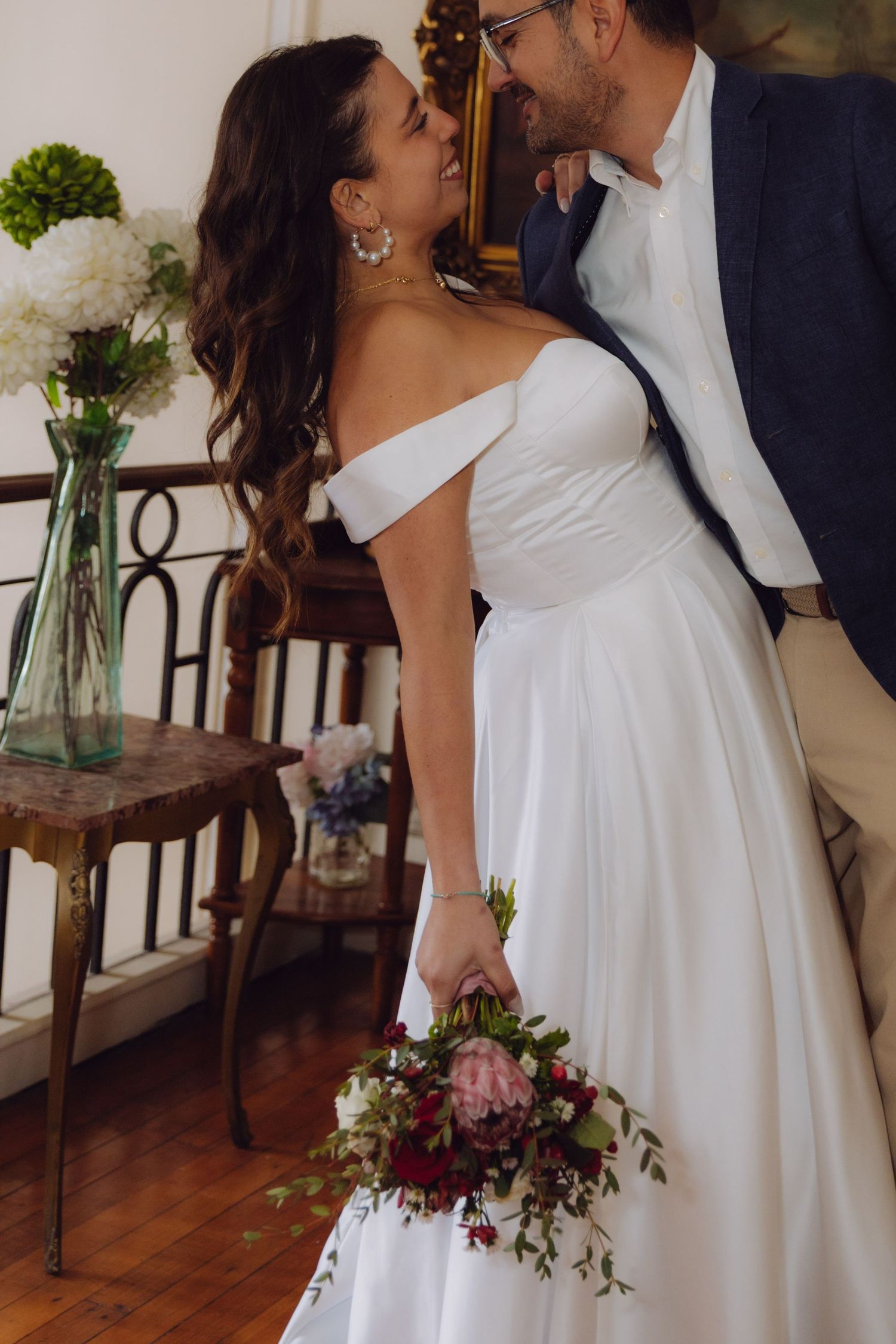 Laughing bride in off-shoulder gown leans into groom beside floral arrangements on wooden table.