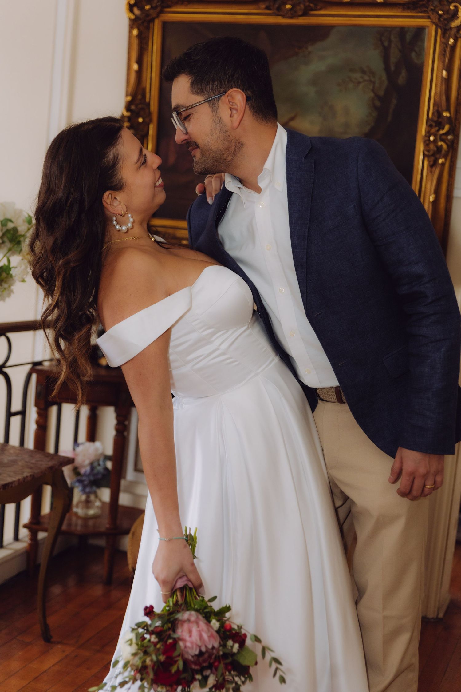 Bride and groom share a romantic smile near an ornate gold-framed painting in a classic interior.