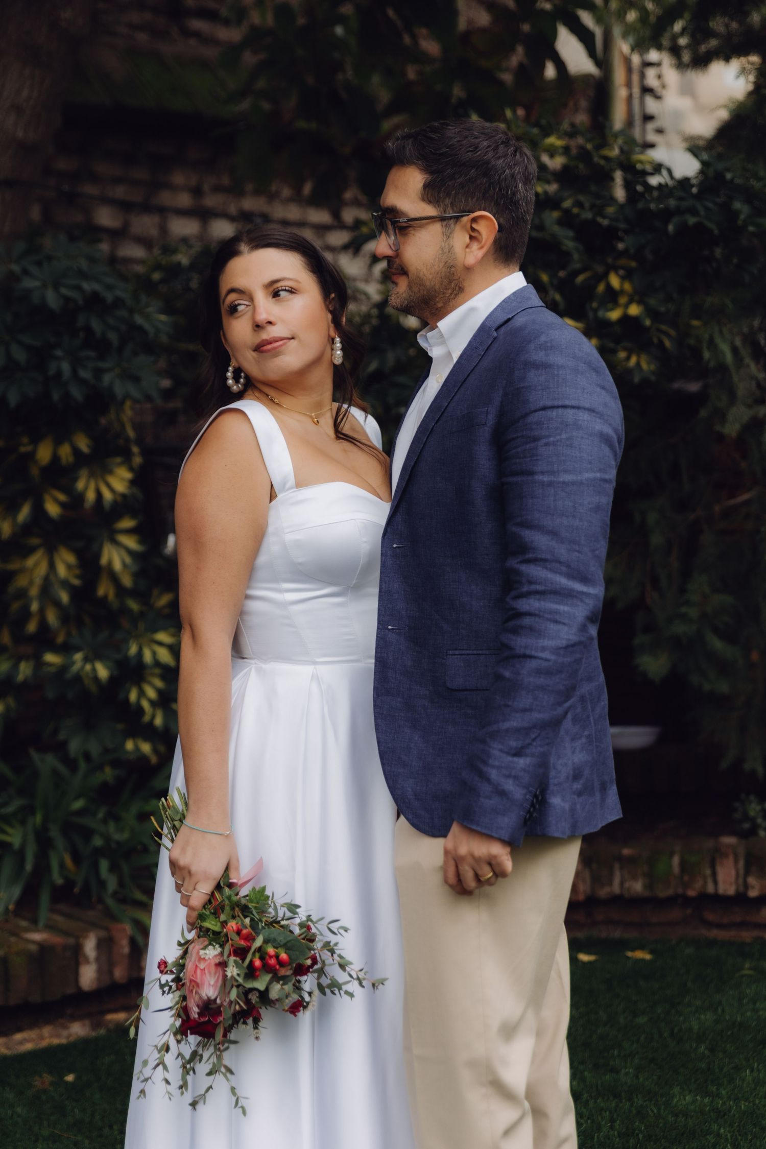 A bride in white dress and groom in blue blazer pose in a lush garden, bride holding a red floral bouquet.