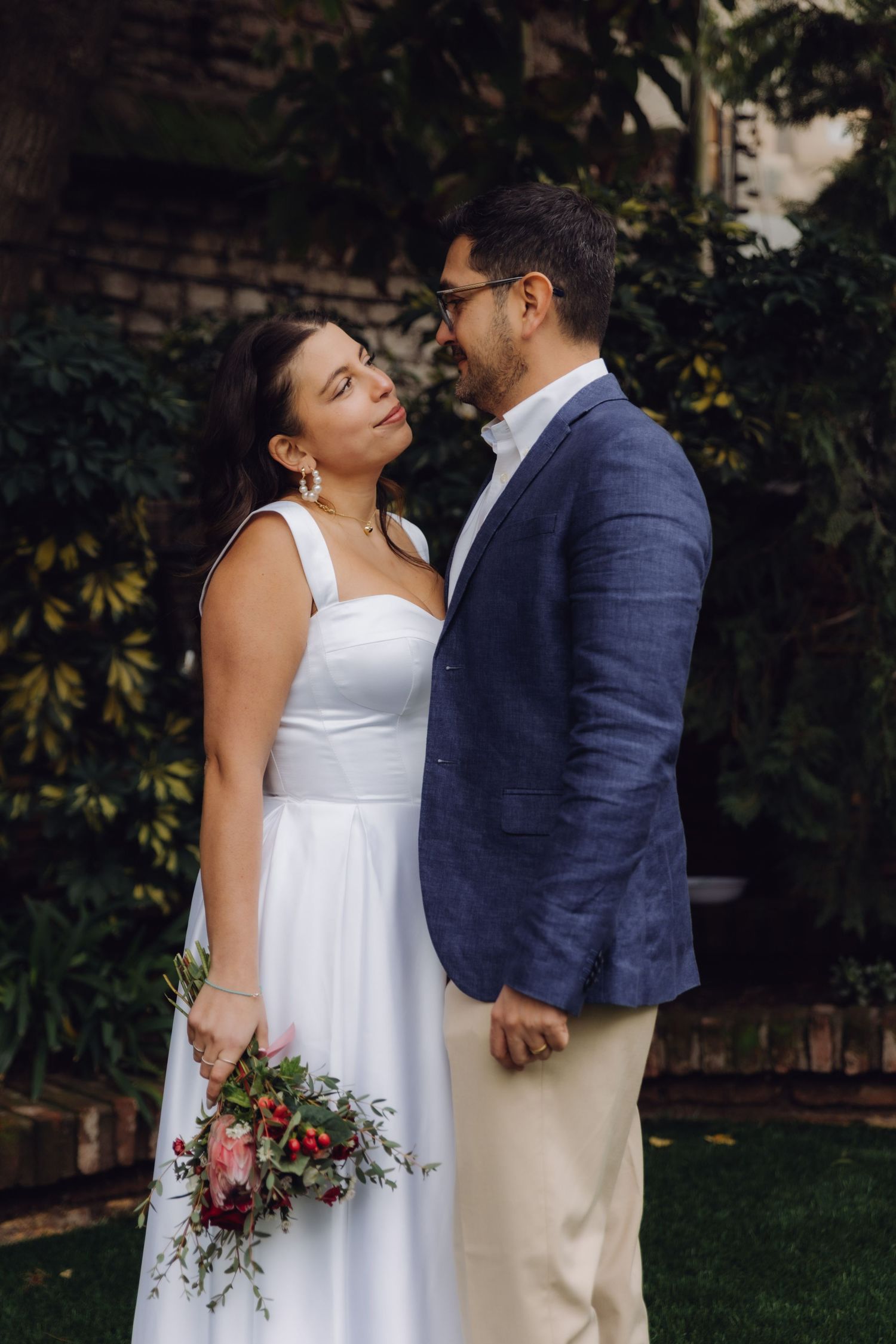A bride and groom gaze into each other's eyes in a garden setting, bride holding a vibrant red bouquet.