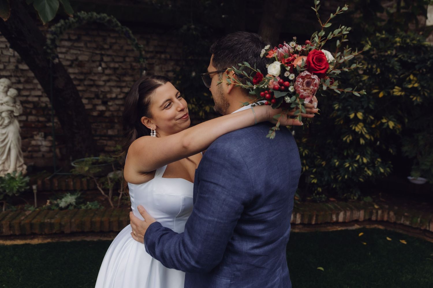 A bride embraces her groom in a garden, holding a red and pink bouquet while smiling up at him lovingly.