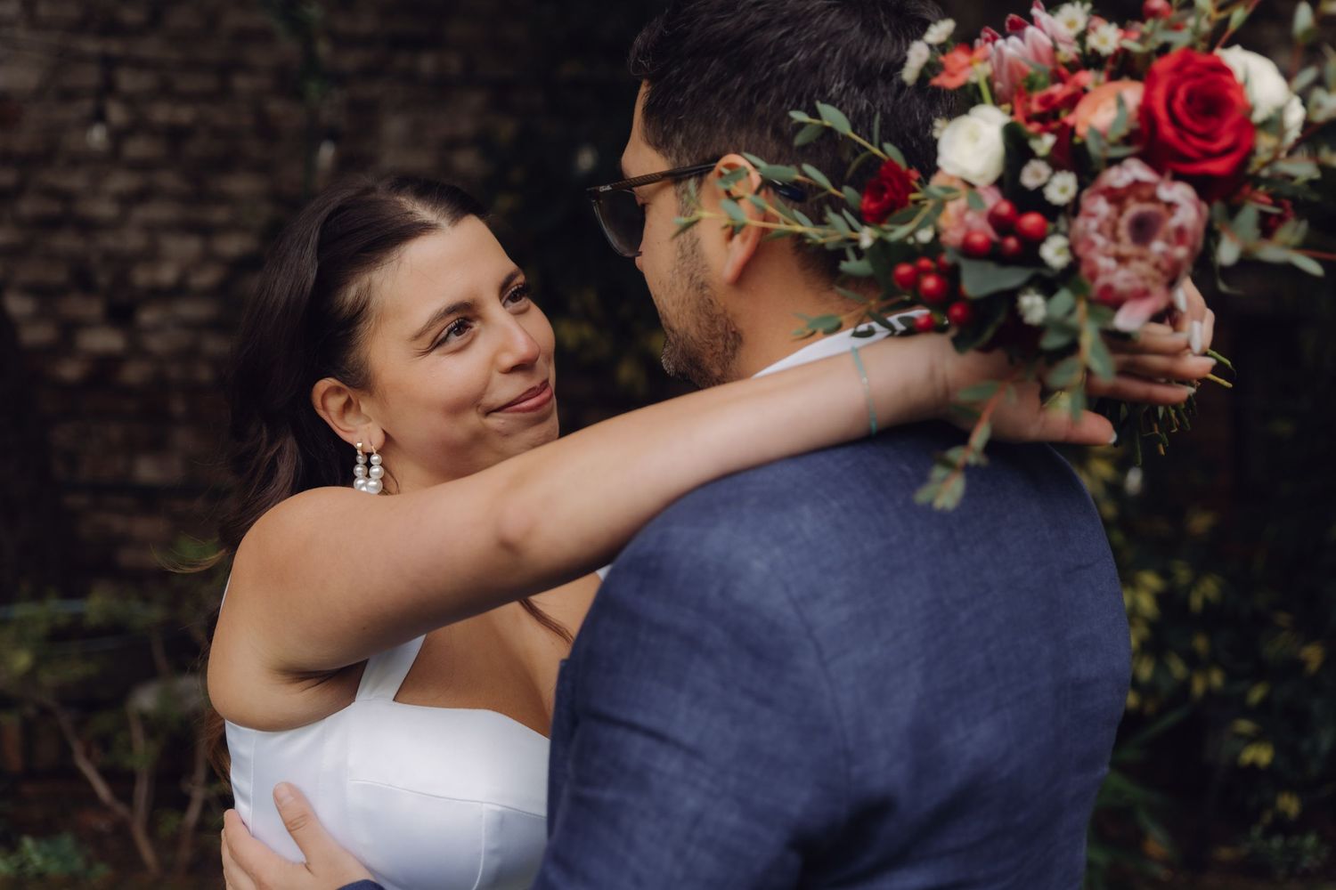 Bride gazes lovingly at groom while holding red and white wedding bouquet outdoors.