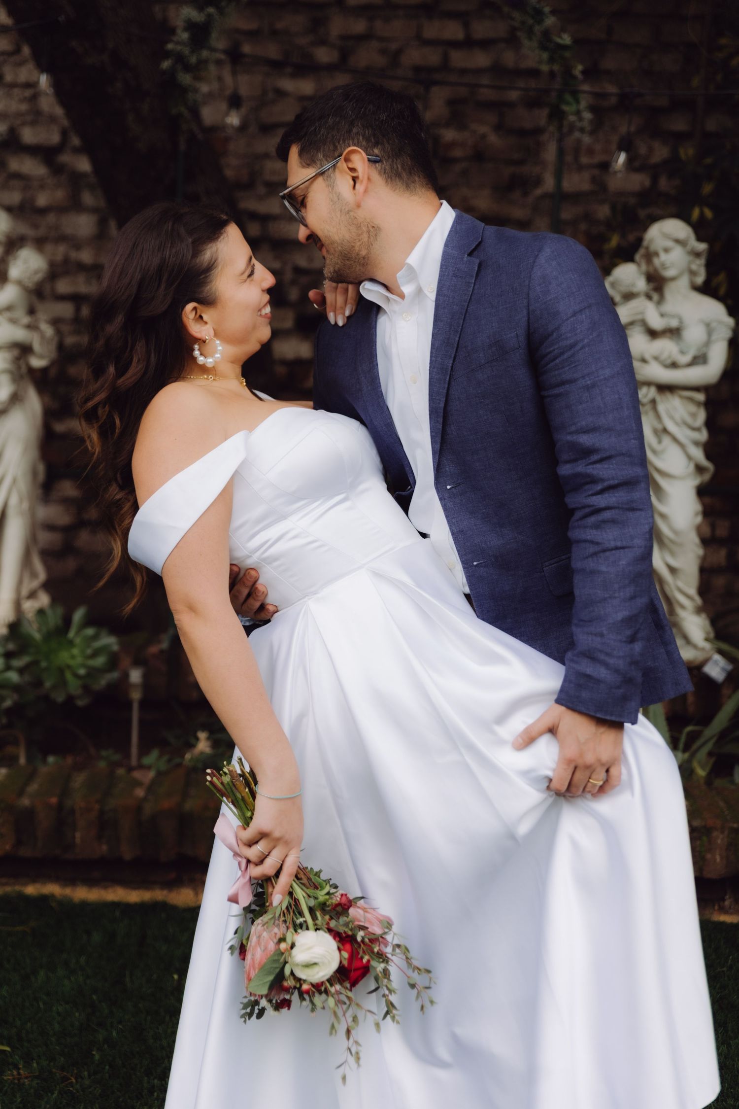 Bride in off-shoulder white gown and groom in blue suit embrace near garden statues.
