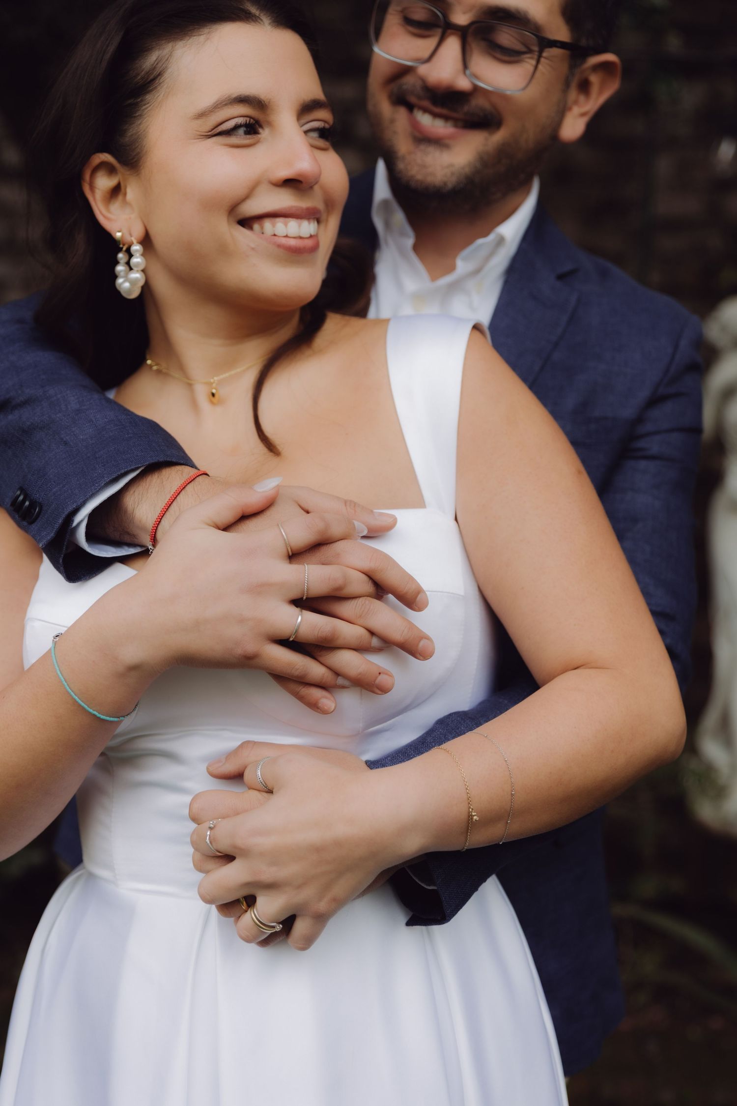 Groom tenderly embraces smiling bride in white dress, hands intertwined, showing wedding rings.