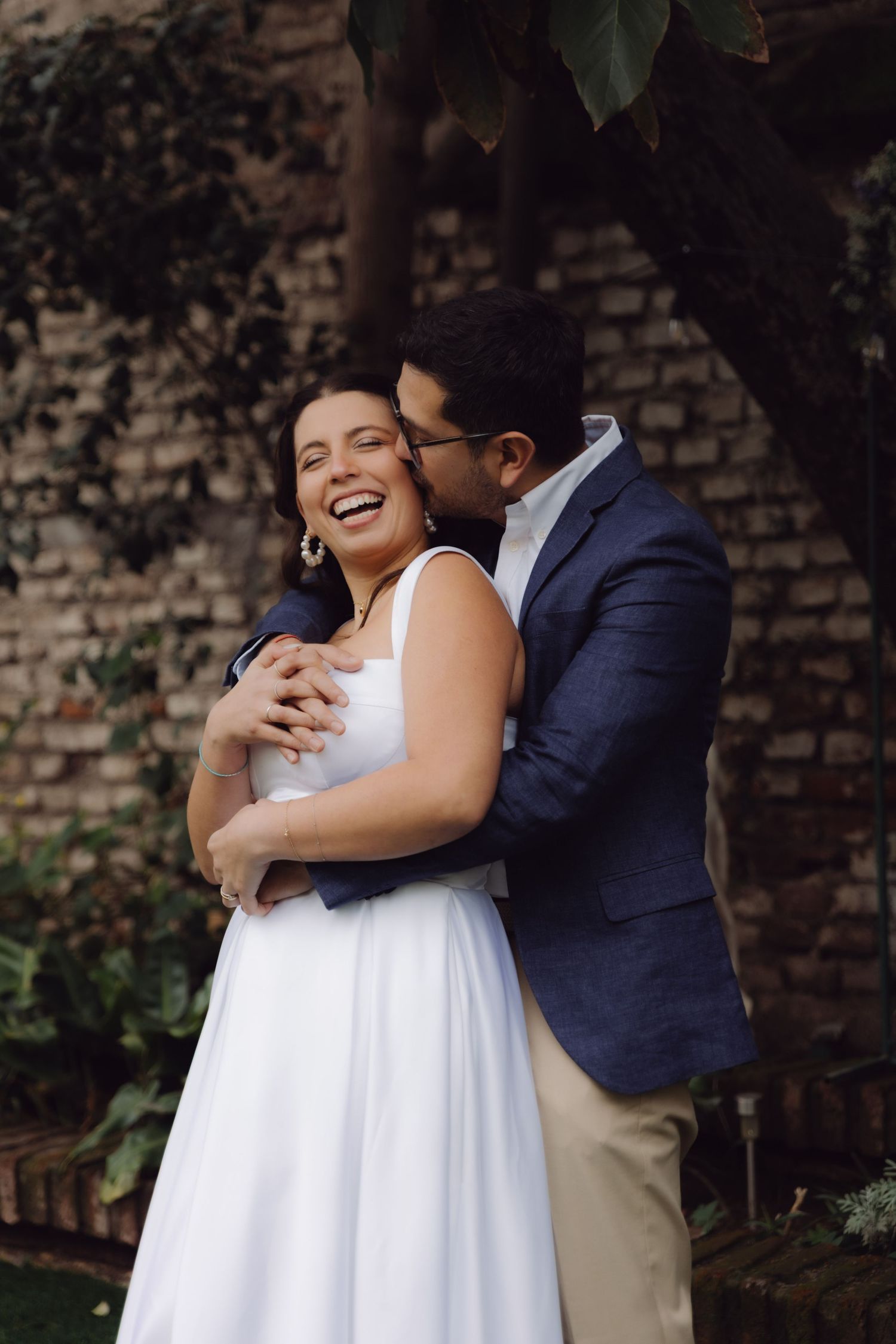 Groom kisses laughing bride from behind in garden setting with brick wall backdrop.