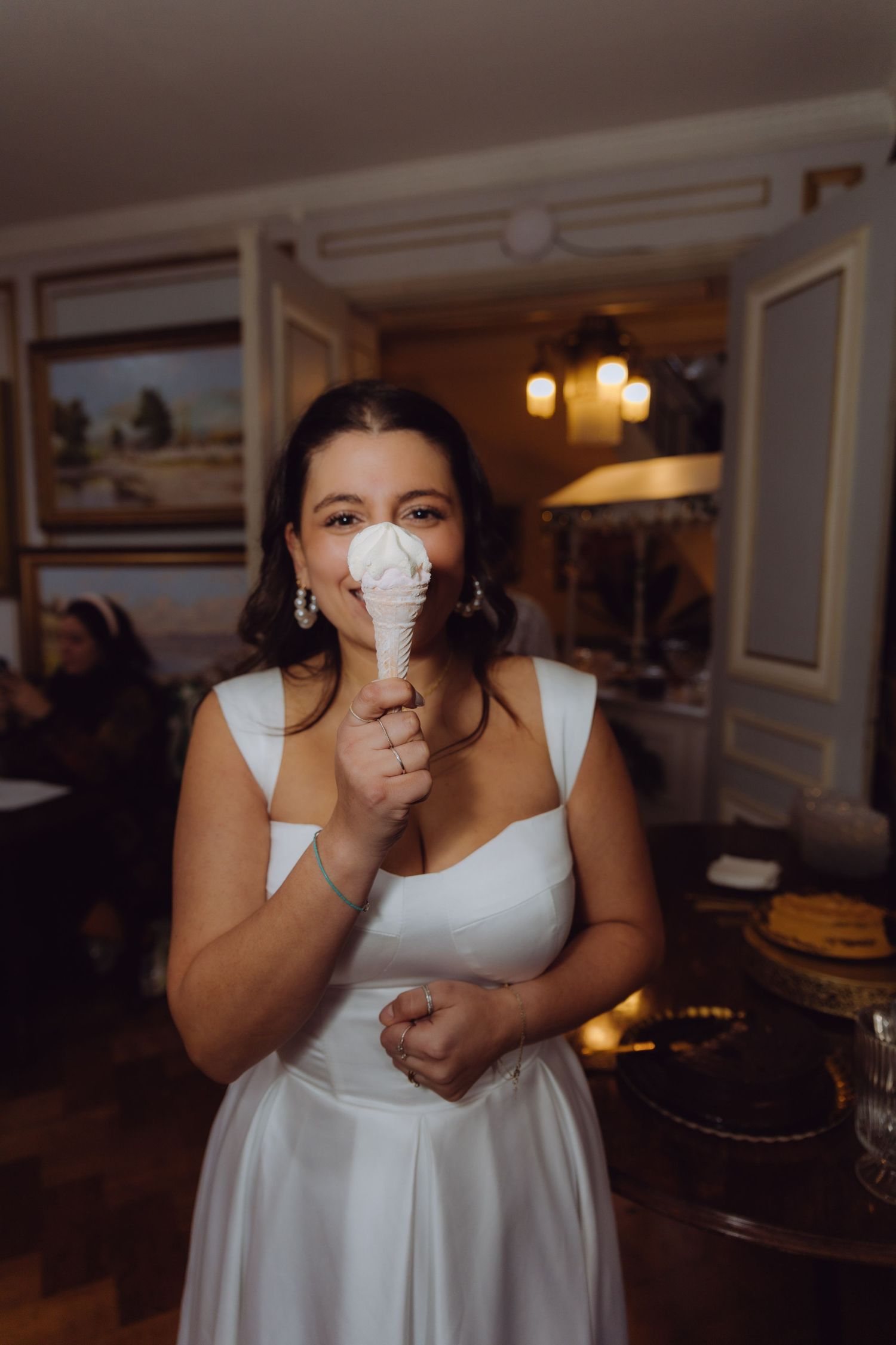 Happy bride in white wedding dress playfully holds ice cream cone in elegant venue.
