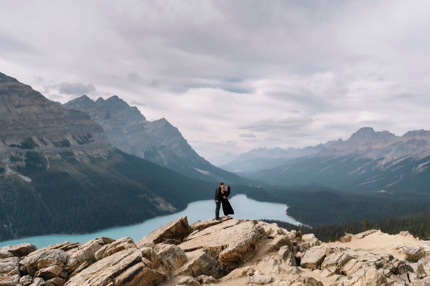 Panoramic mountain landscape view of a proposal couple at Peyto Lake in Banff National Park with dramatic cloudy skies and turquoise waters.