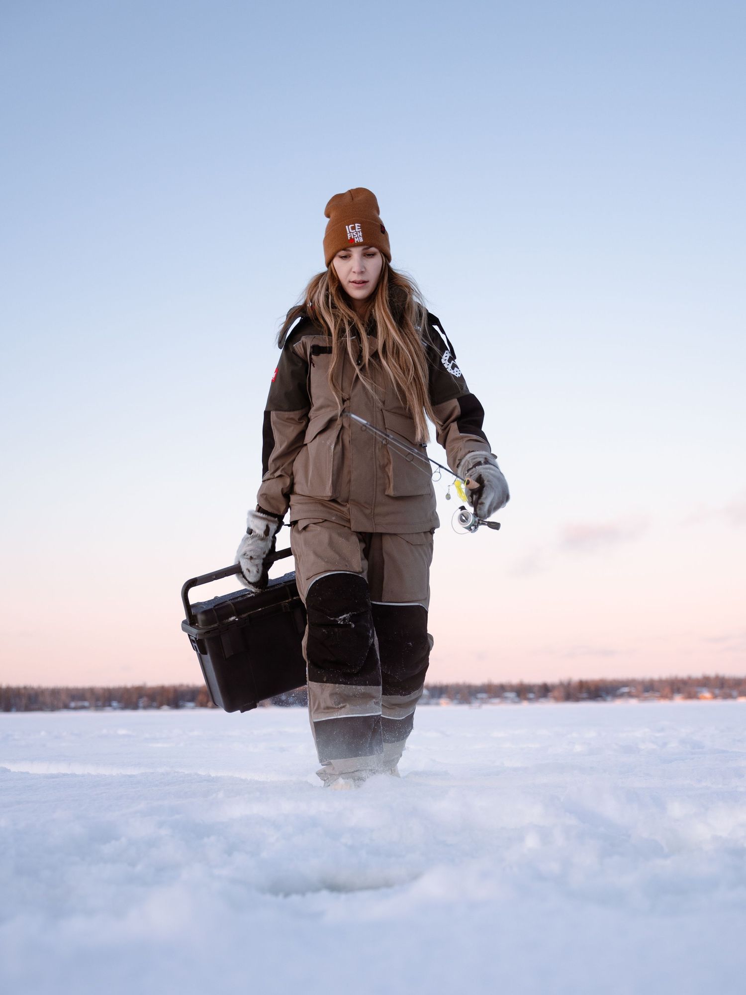 Winter angler walking across frozen lake carrying ice fishing gear at sunset.