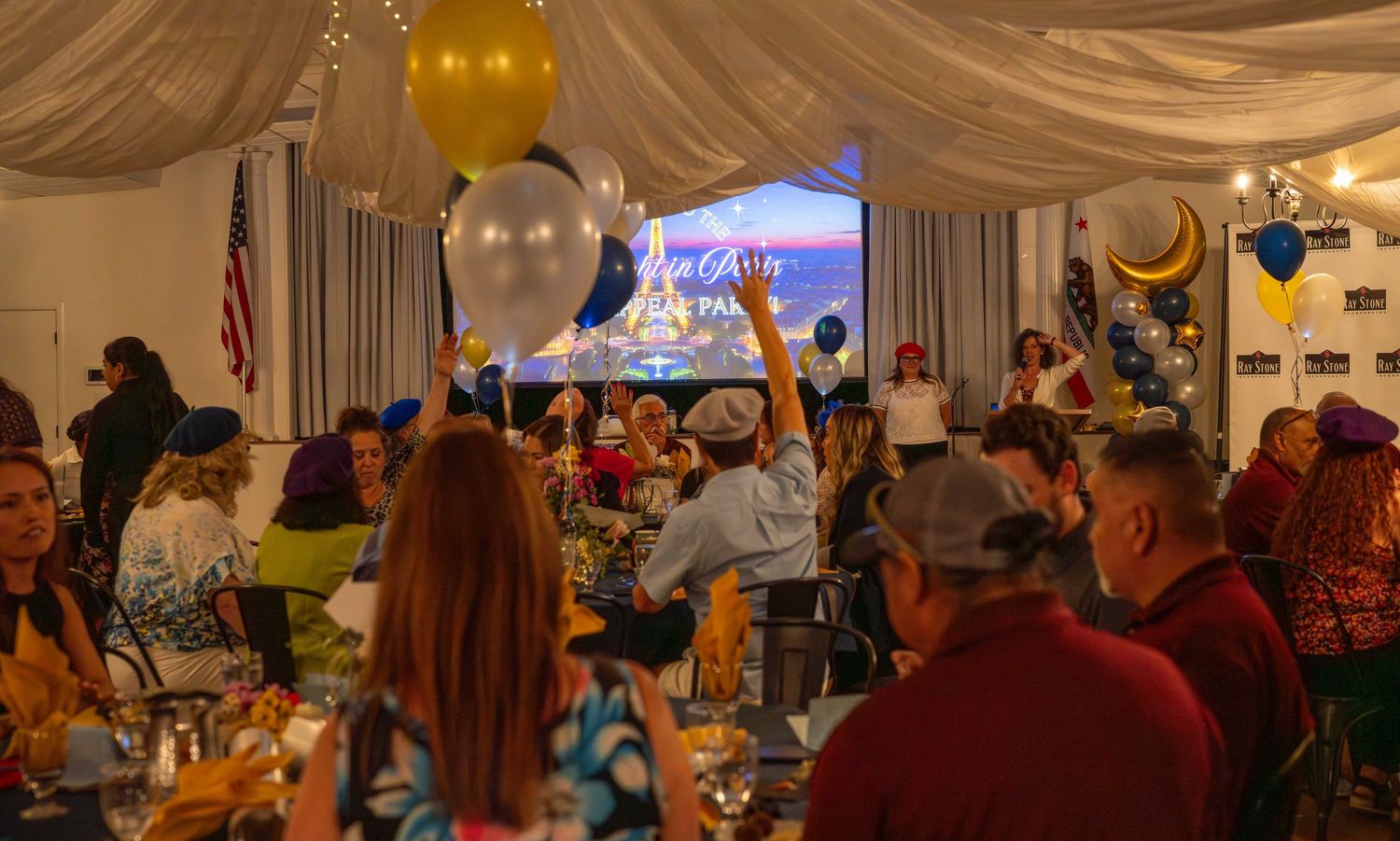 A crowded indoor celebration event with balloons and decorations featuring people seated at tables under draped fabric ceiling.