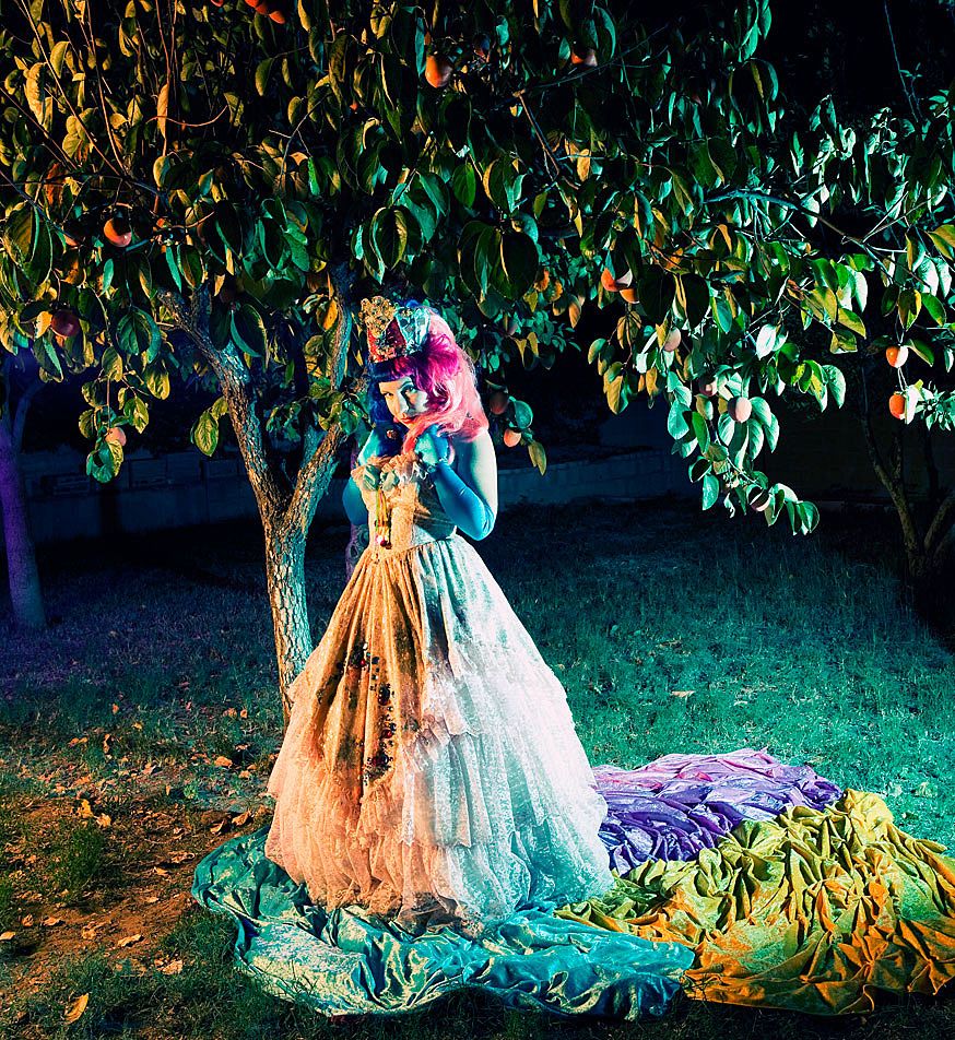 Two elegant ball gowns with full skirts and romantic styling sit beneath a tree at twilight with twinkling lights.