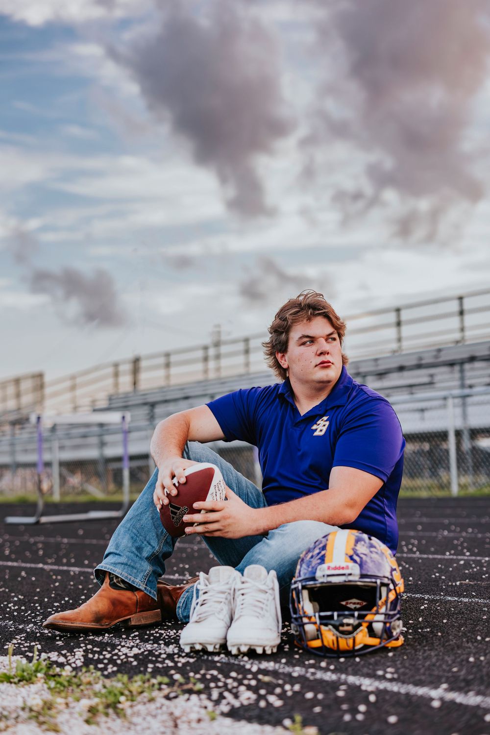 Football player in blue jersey sits with ball and helmet on track at sunset.