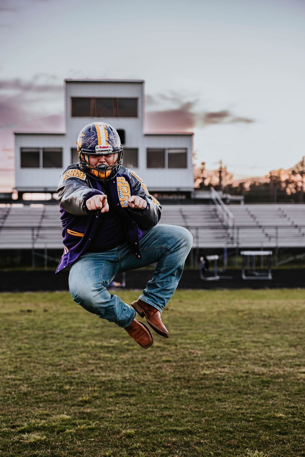 Football player in helmet and jeans jumps in mid-air on field at sunset.