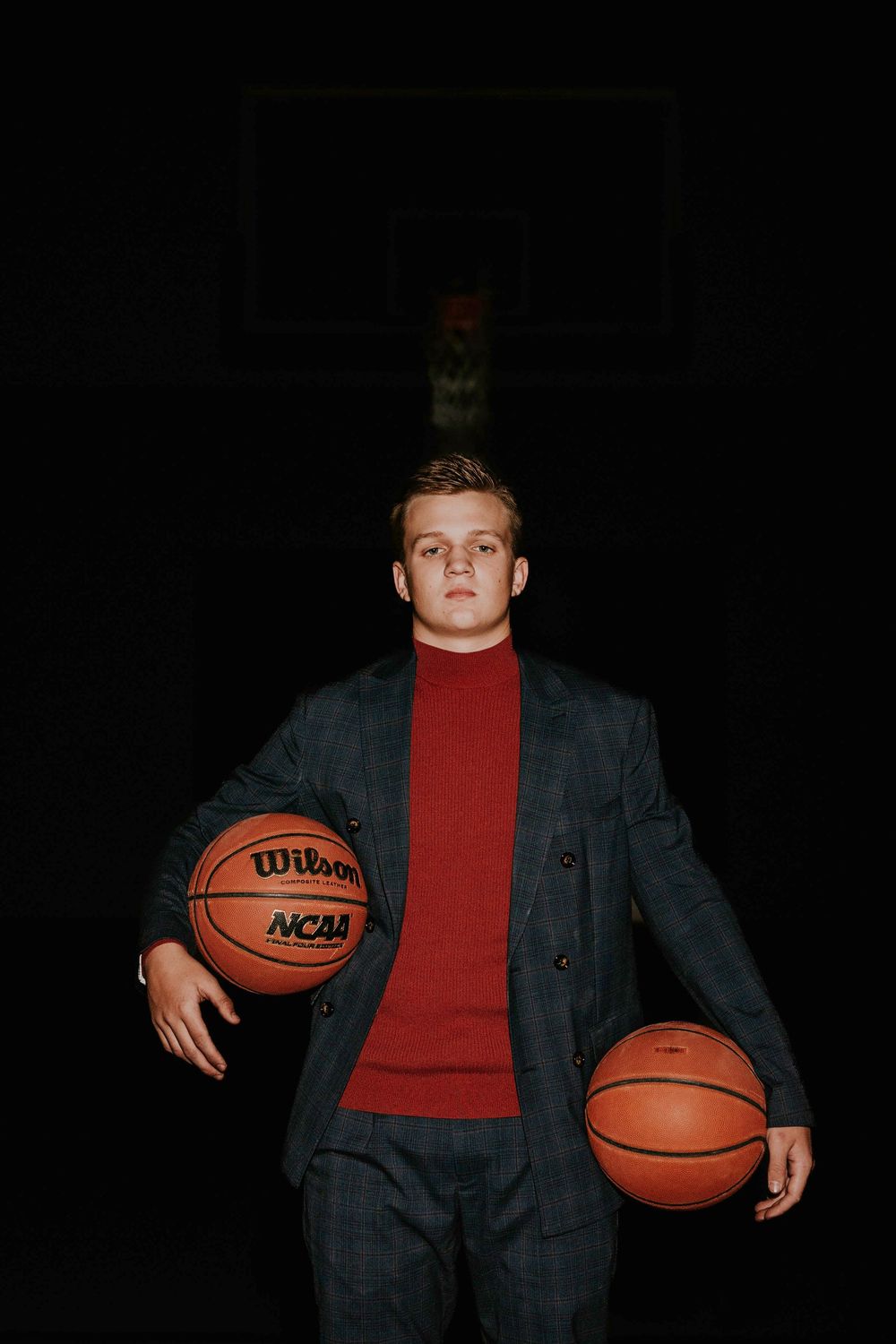 Basketball player in dark suit jacket and red shirt poses with basketball in studio setting.