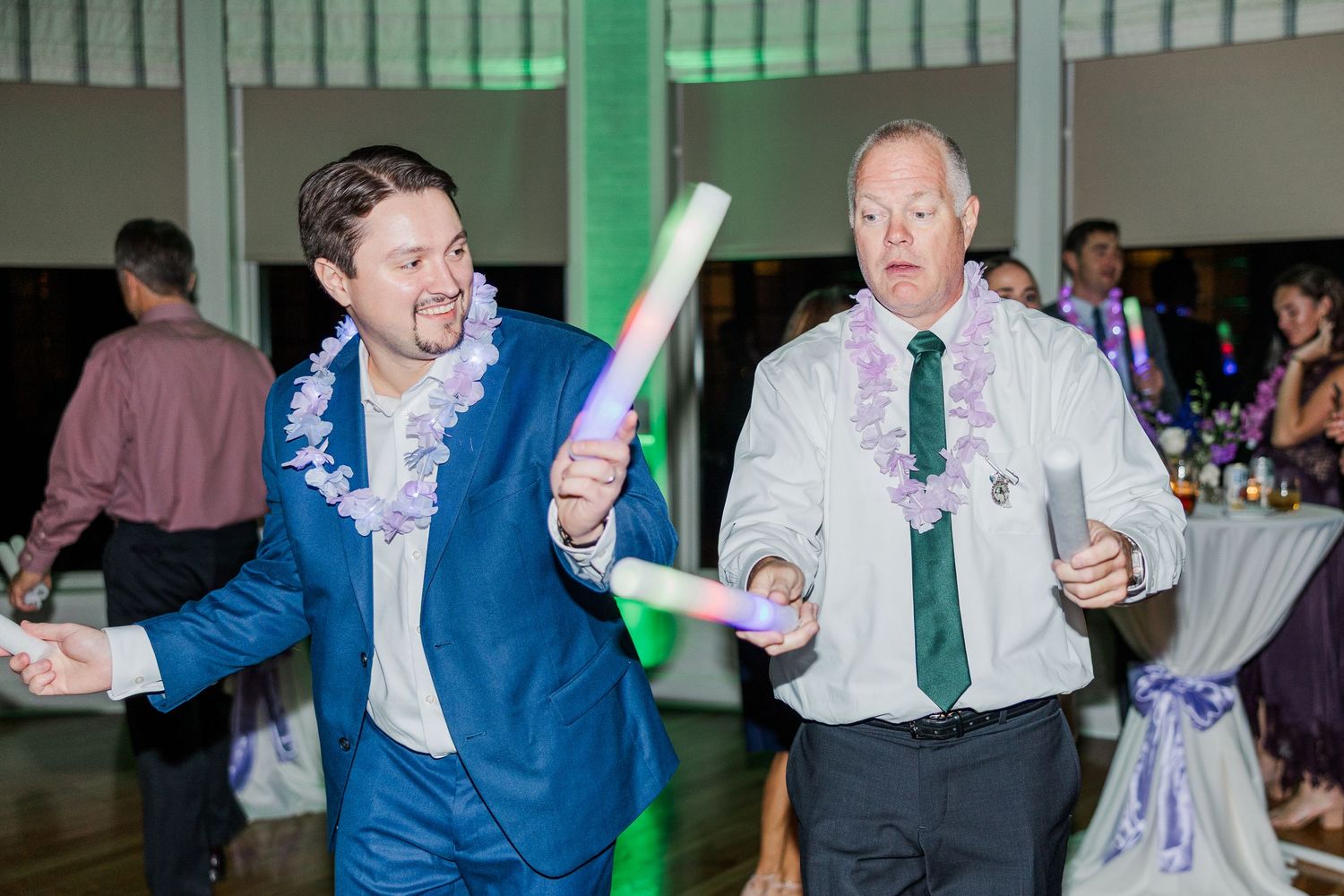 Two wedding guests in suits dance energetically at a reception with one holding a glow stick and wearing a floral lei.